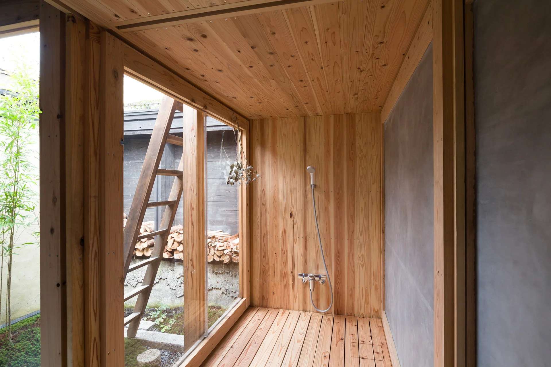 A minimalist shower room features light natural wood paneling on the walls, ceiling, and slatted floor alongside a smooth grey concrete wall.