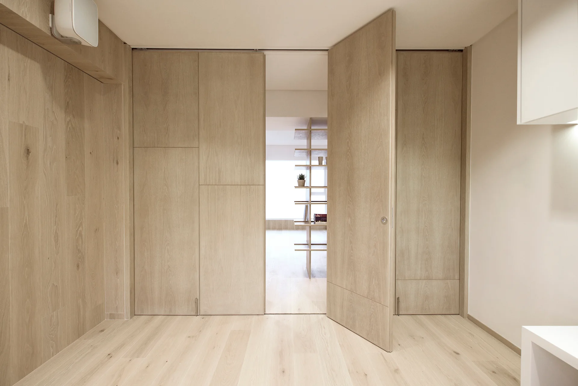 A light wood paneled hallway features floor-to-ceiling pivoting doors that open to reveal an adjacent room with open wooden shelving.