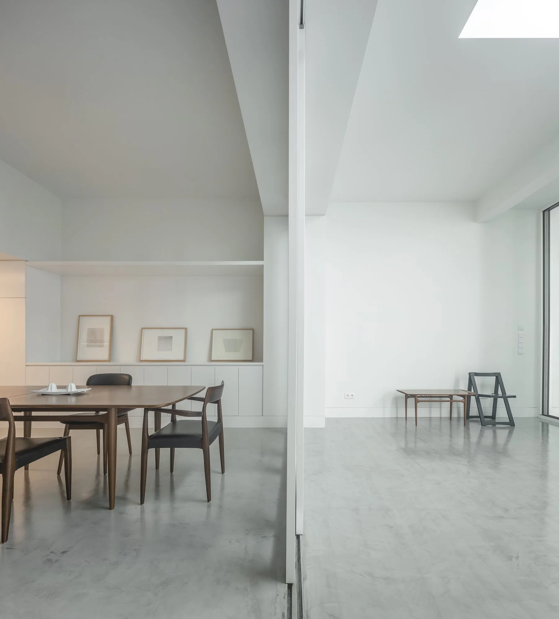 A minimalist dining room featuring a wooden table and chairs on concrete floors next to a white built-in storage unit and a partition wall.