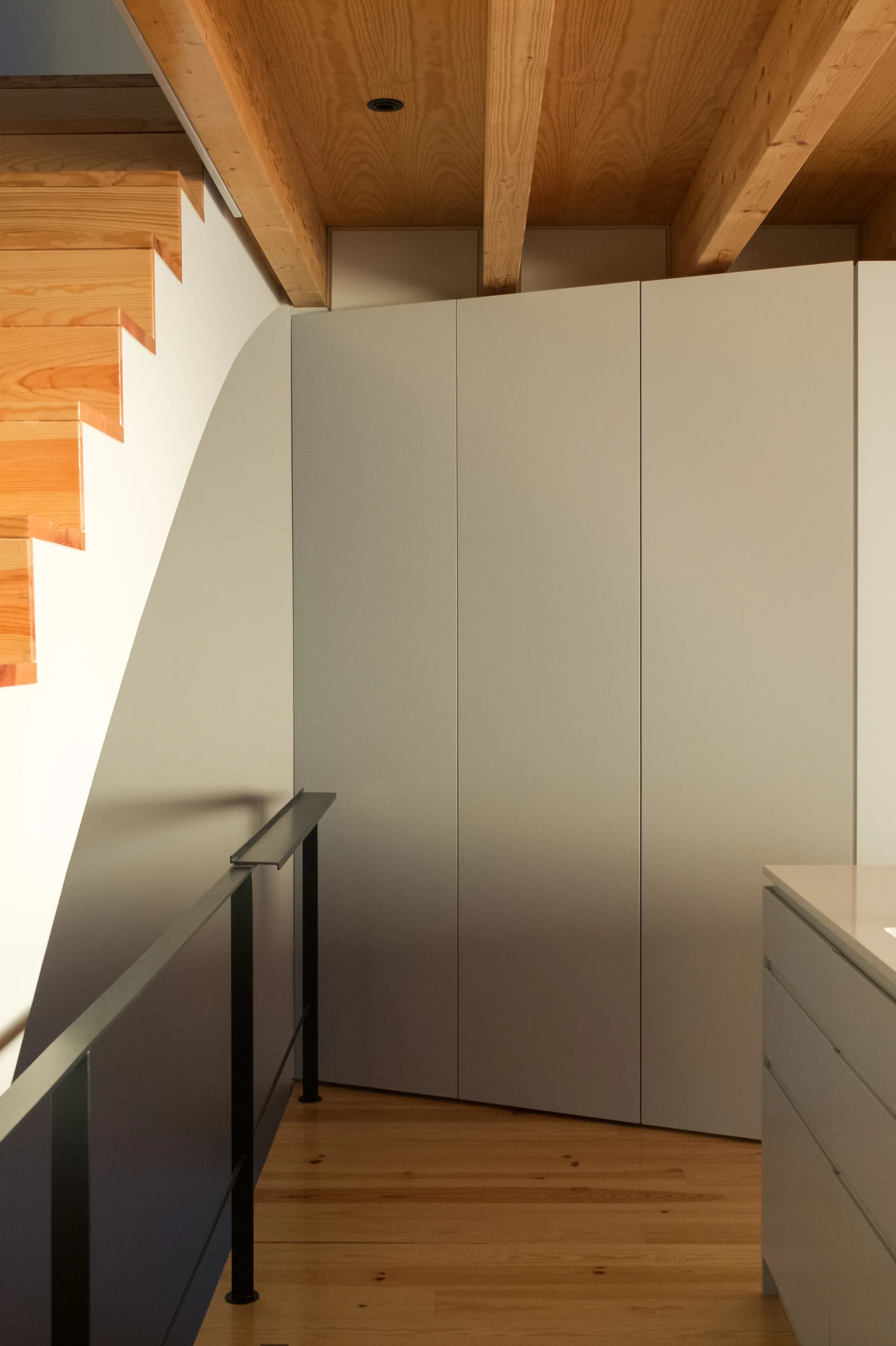A minimalist light gray built-in cabinet wall is integrated into a staircase landing with wood floors and exposed timber ceiling beams.