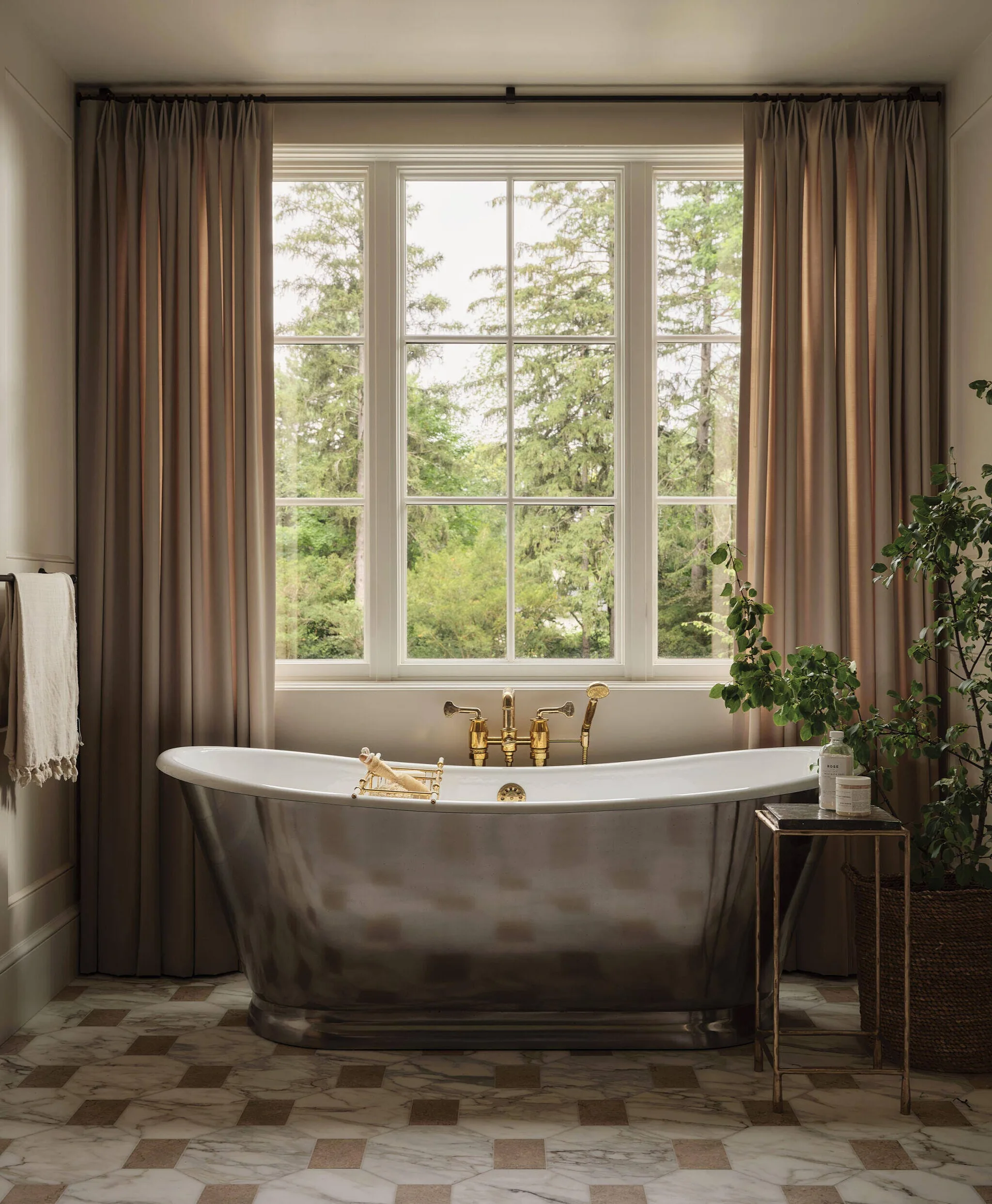 A silver metallic freestanding bathtub with gold fixtures sits in front of a window flanked by taupe curtains on a checkerboard marble floor.