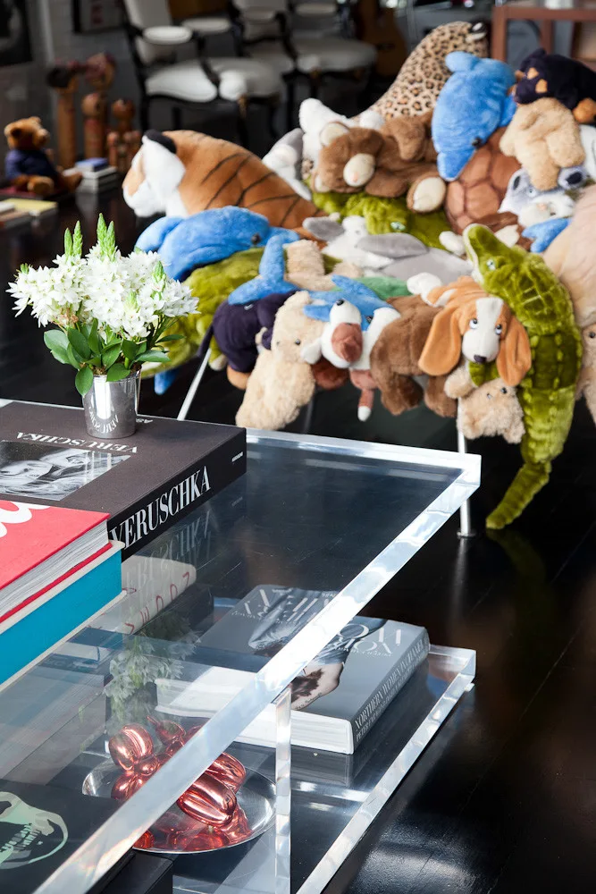 A clear acrylic coffee table displaying stacked books and flowers sits on dark floors in front of a sofa piled high with stuffed animals.