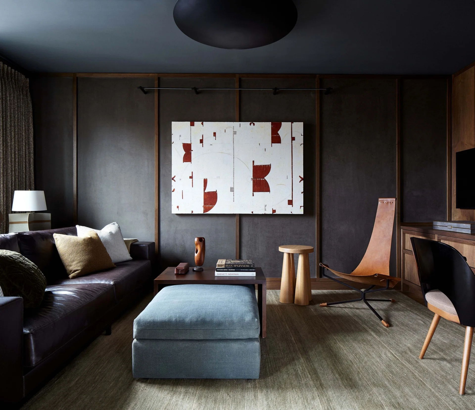 A moody living room features dark wood wall paneling, a dark leather sofa, a textured grey-blue ottoman, and a tan leather sling chair.