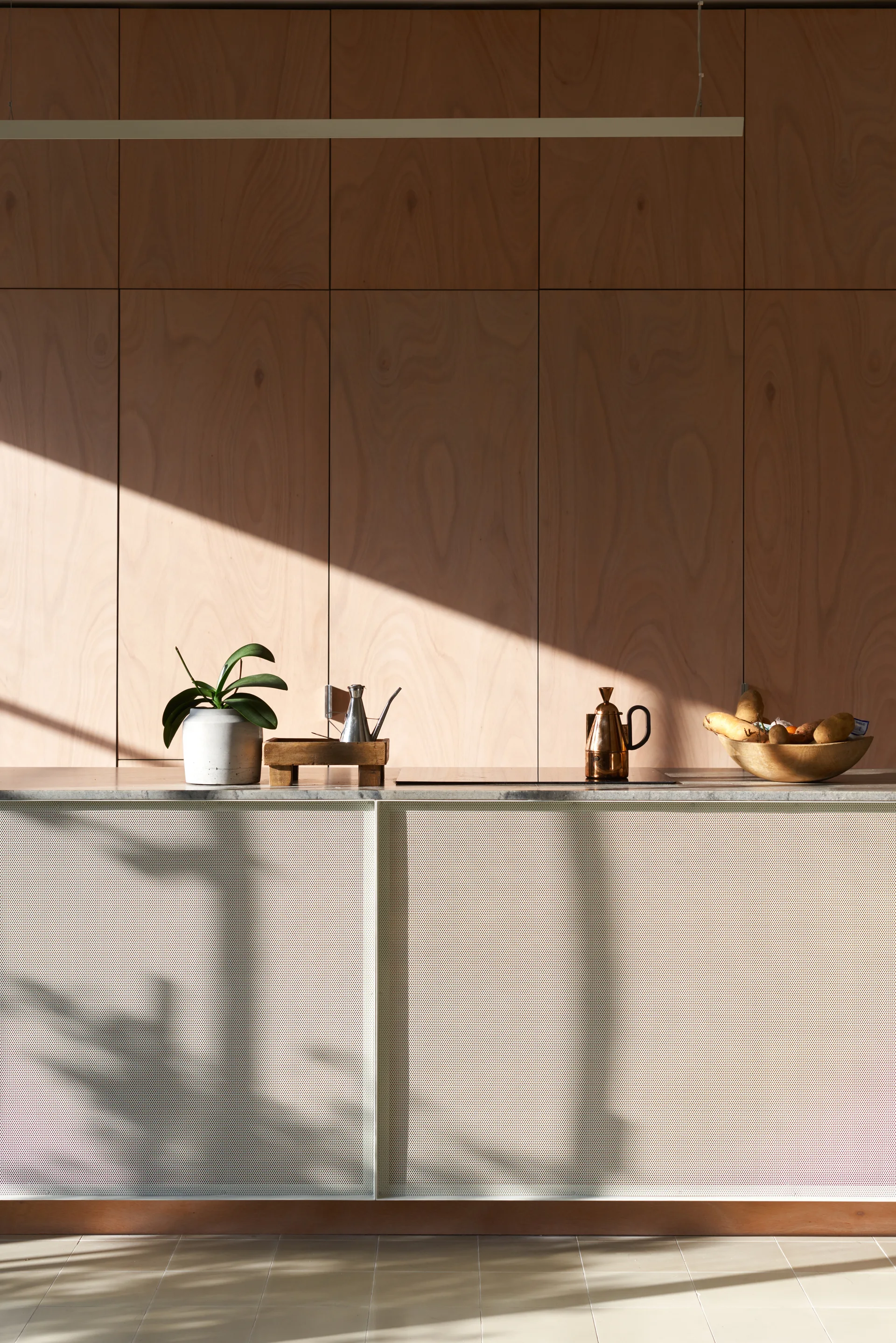 A modern kitchen island with a perforated metal front and stone countertop sits before a warm plywood paneled wall in natural light.