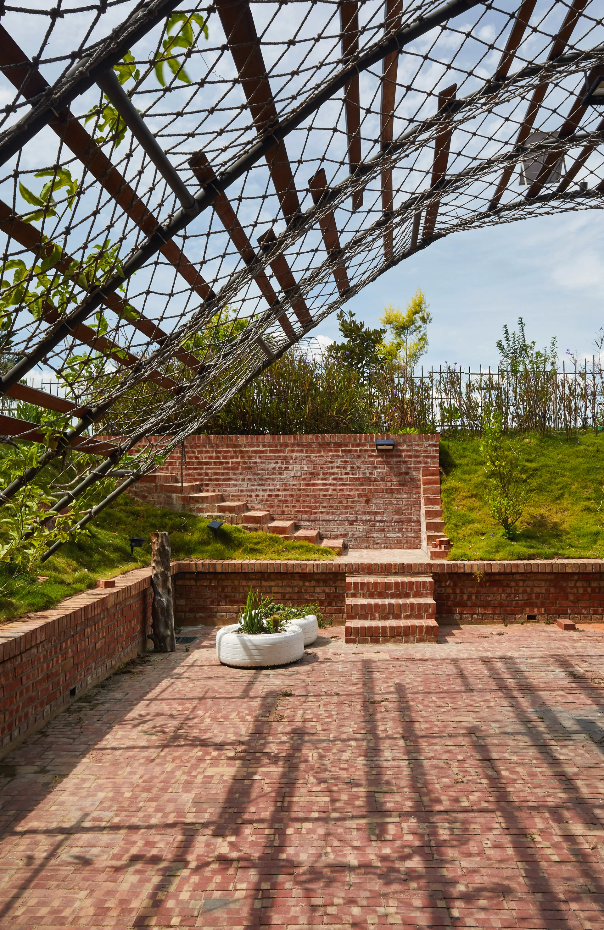 A red brick terrace features a tiered brick staircase built into a grassy slope underneath a large curved wooden trellis with netting.