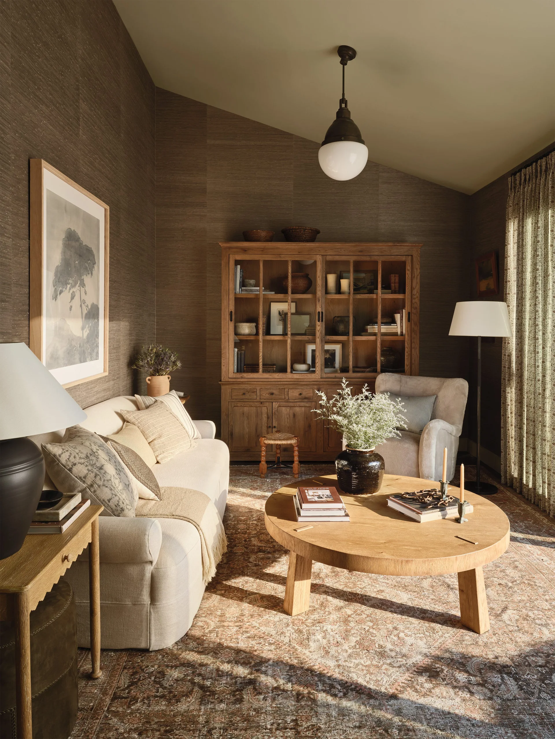 A living room features textured brown wallpaper, a cream-colored sofa, a round wooden coffee table, and a tall wooden display cabinet.