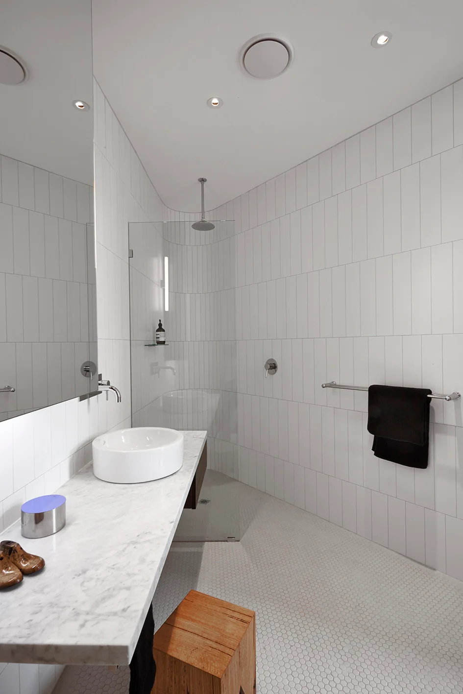 A modern white bathroom features a floating marble vanity with a vessel sink, vertical wall tiles, a penny tile floor, and a curved glass shower screen.