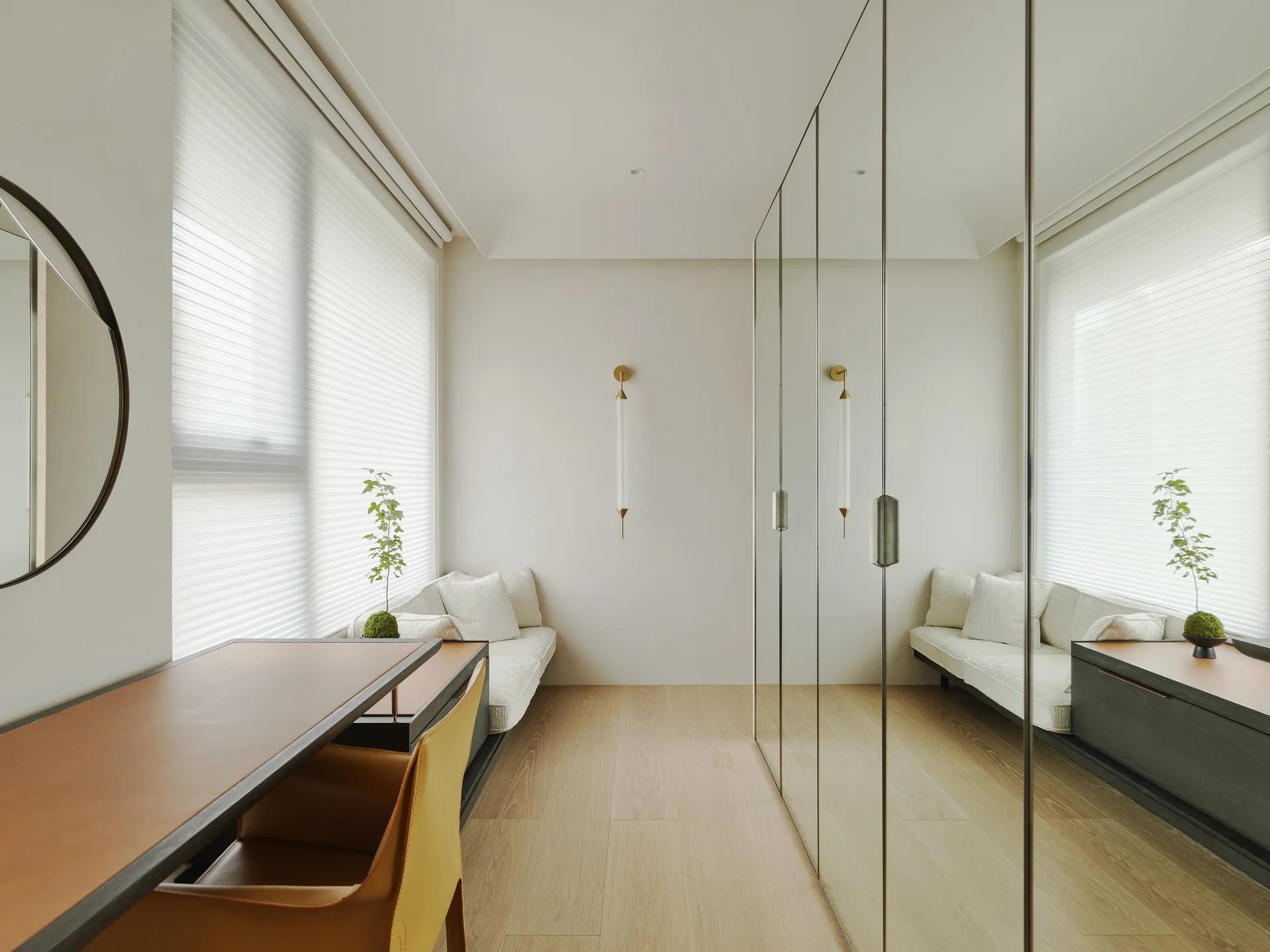 A modern dressing room features a floor-to-ceiling mirrored wardrobe, a built-in window bench, and a desk with a tan leather top.