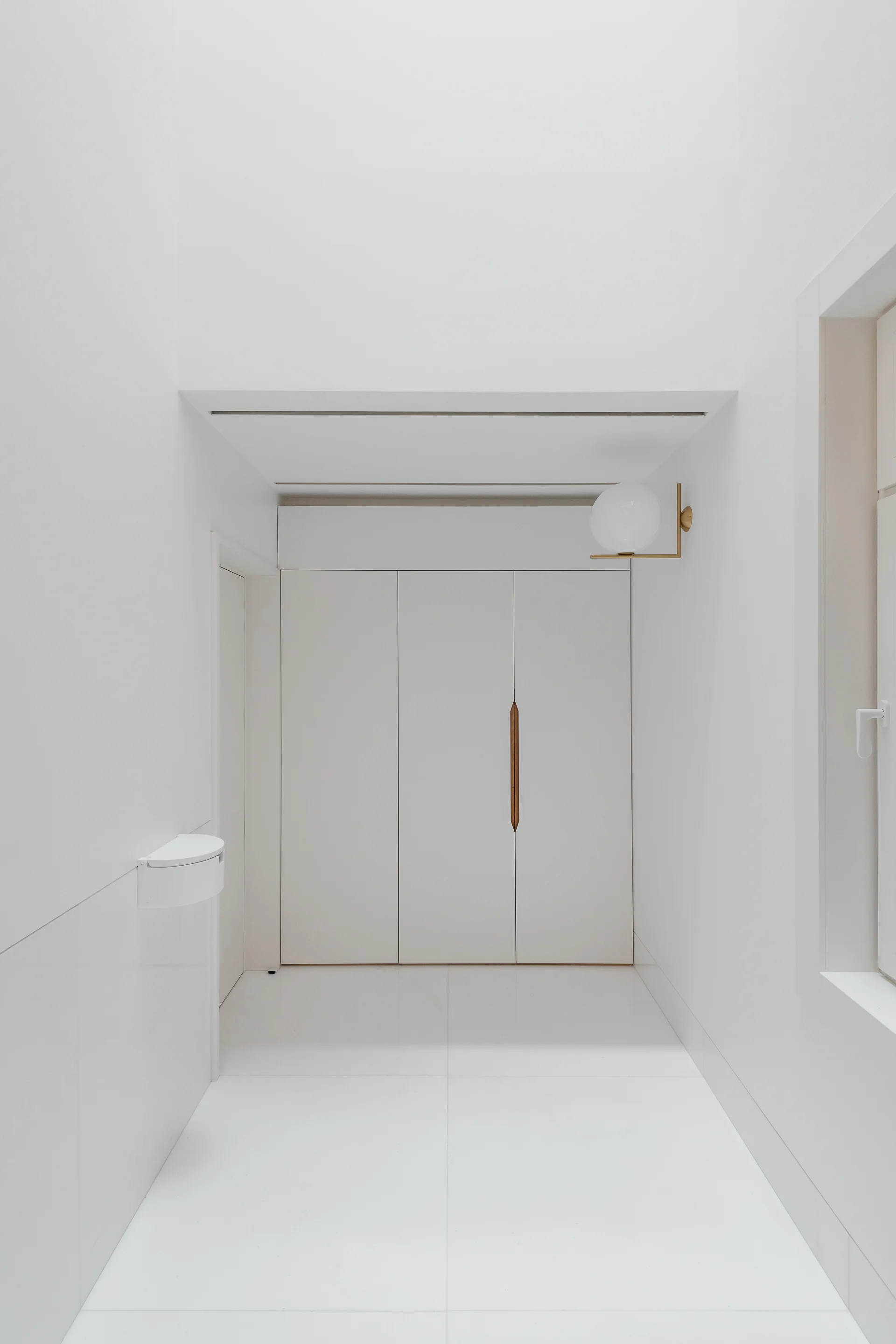 A minimalist white hallway featuring integrated floor-to-ceiling closets with a wooden handle, large floor tiles, and a brass globe wall sconce.