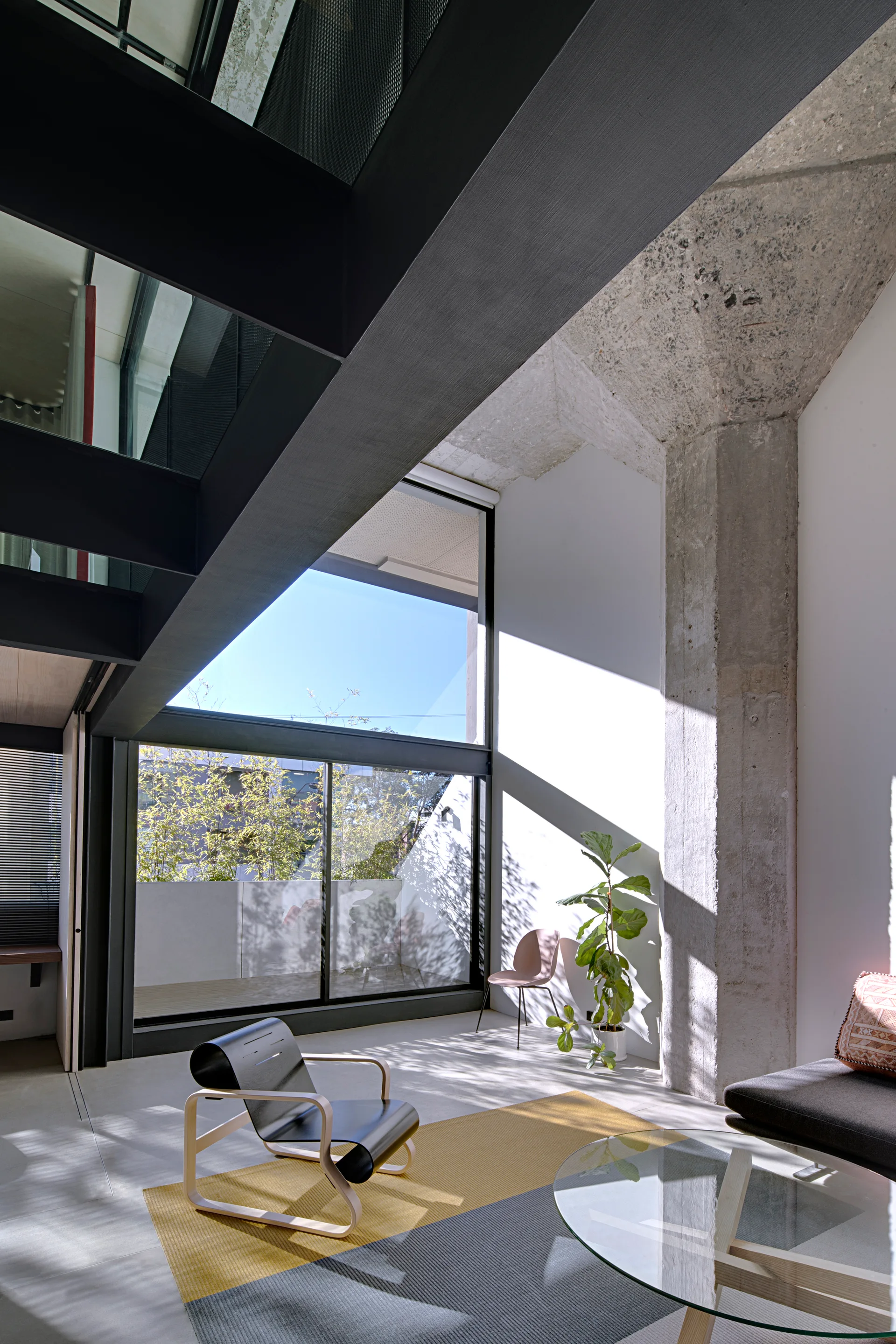 A modern living area featuring a rough concrete pillar, dark metal beams, a curved black lounge chair, and a color-blocked yellow and grey rug.