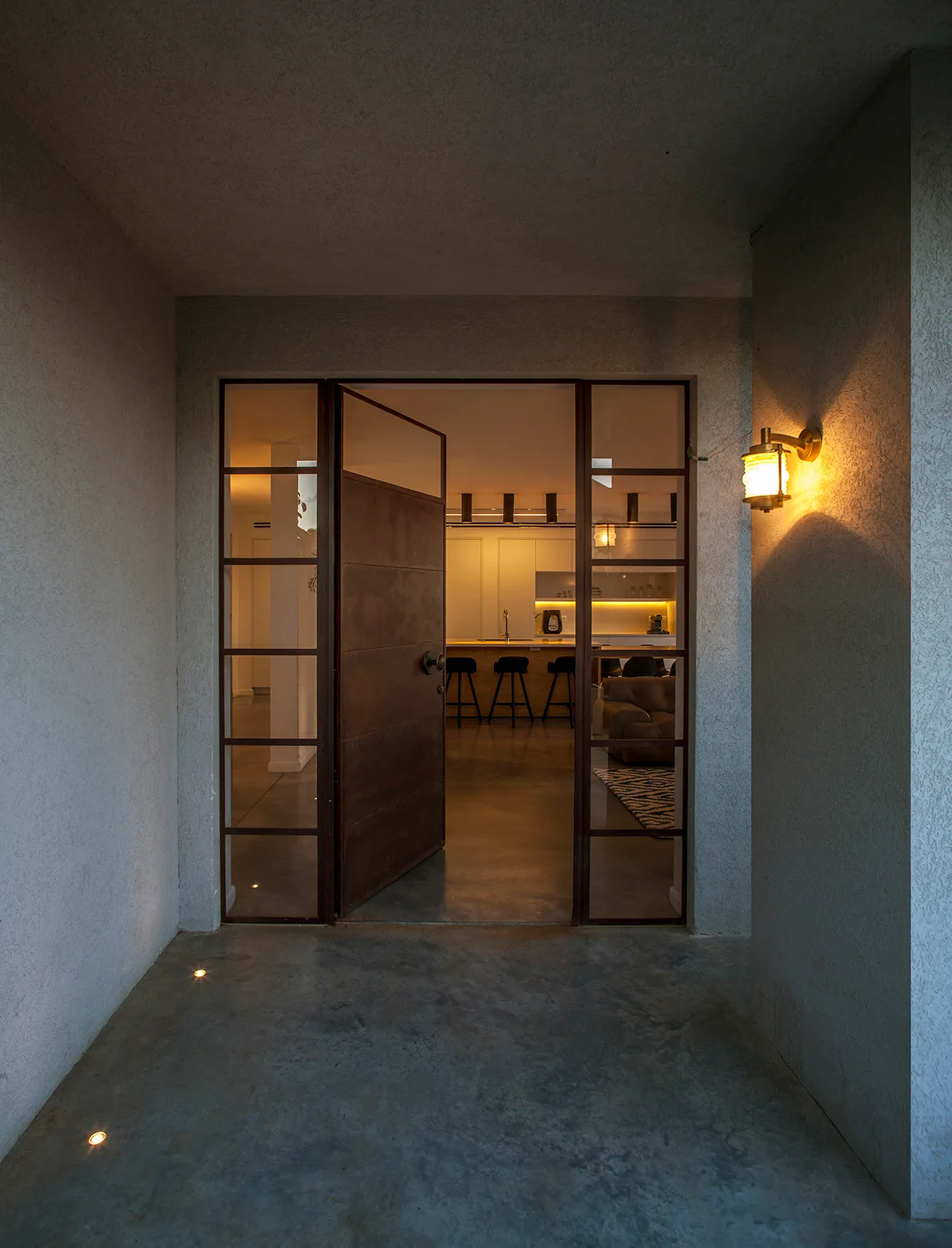 An exterior stucco entryway features a large metal pivot door opening into a modern kitchen with a lighted island and barstools.