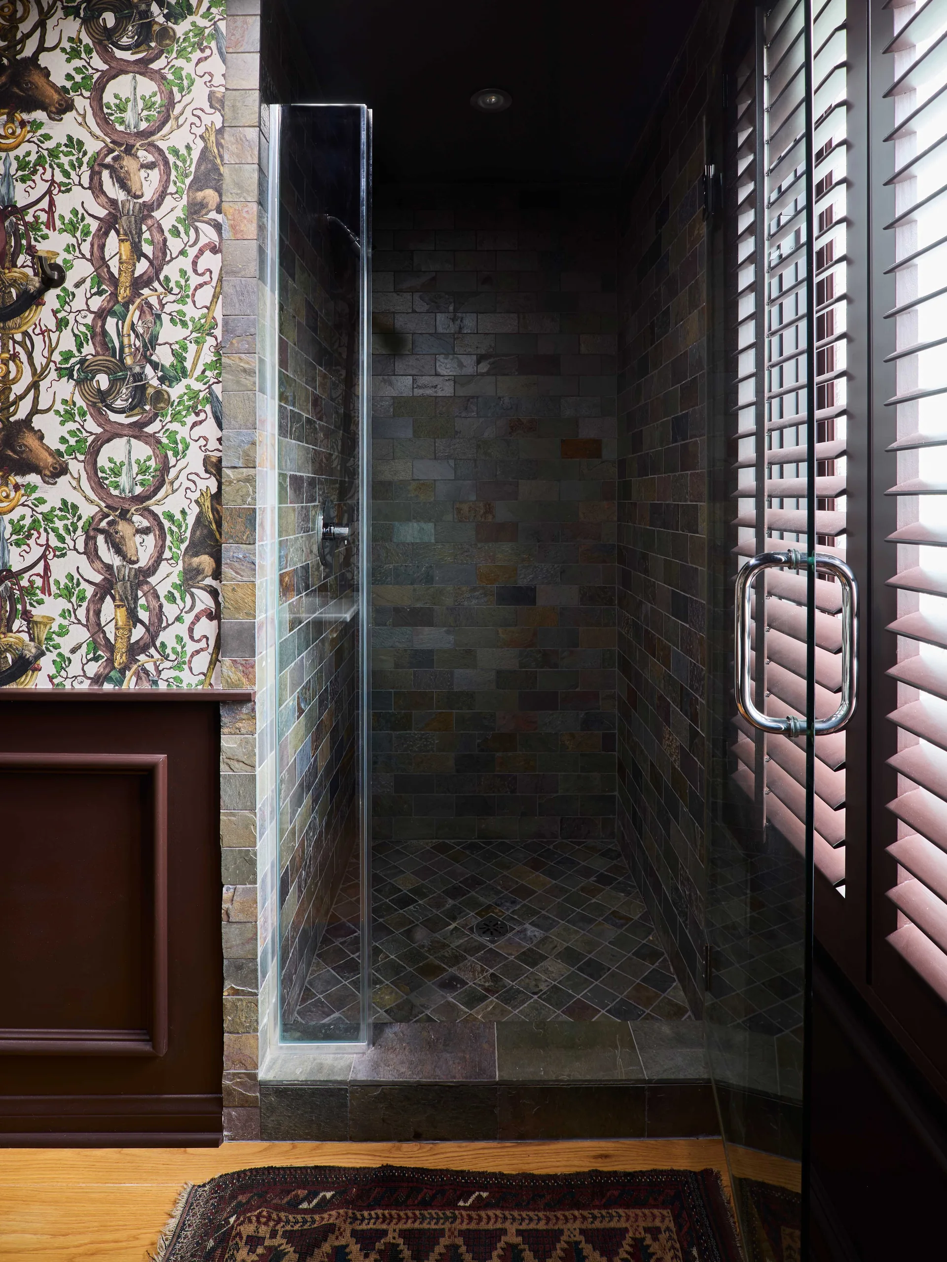 A walk-in shower with earth-toned slate tiles next to a wall with dark wood wainscoting and green and brown animal-motif wallpaper.