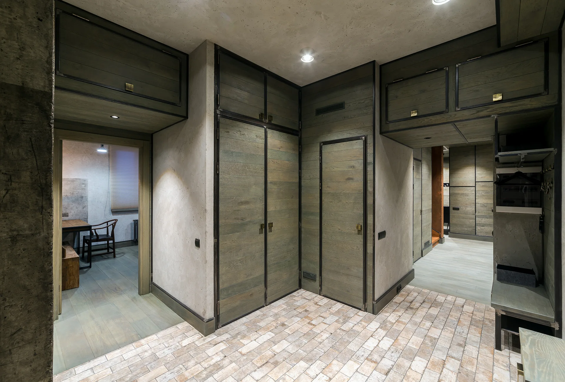 An industrial entryway features built-in weathered wood cabinets with dark metal trim, set against concrete walls and brick floor tiling.