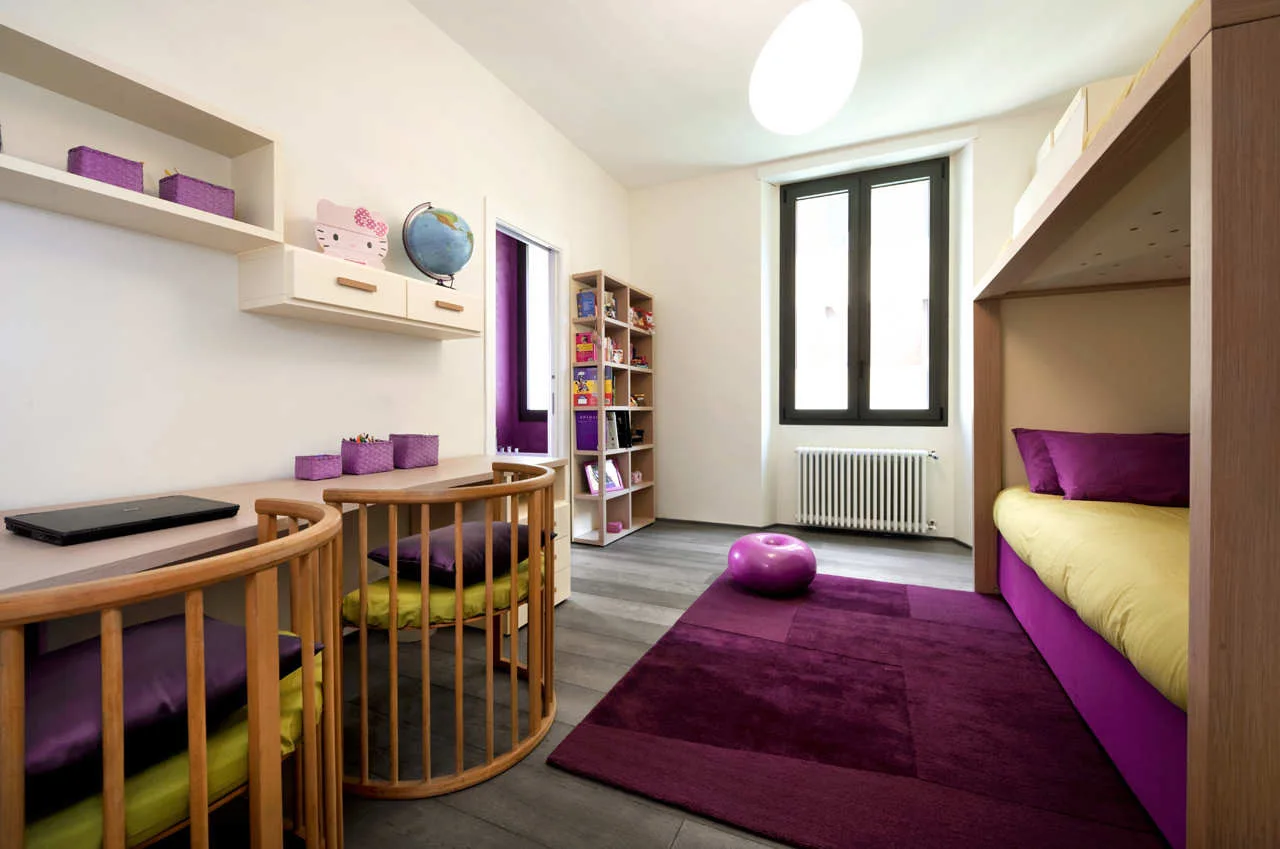 A kids bedroom featuring a wooden bunk bed, a long study desk with two curved spindle chairs, and a large purple rug on grey flooring.
