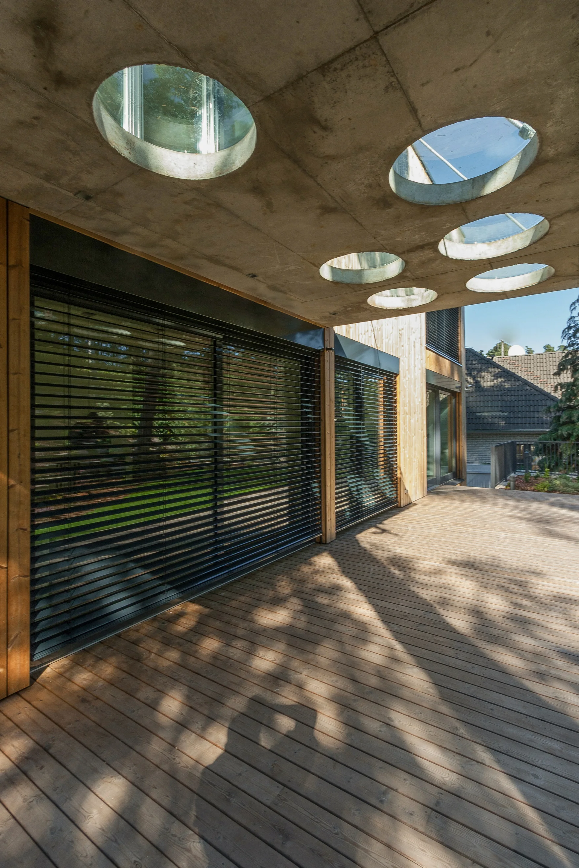 A wooden deck terrace beneath a concrete overhang featuring circular glass skylights alongside large windows with black horizontal blinds.