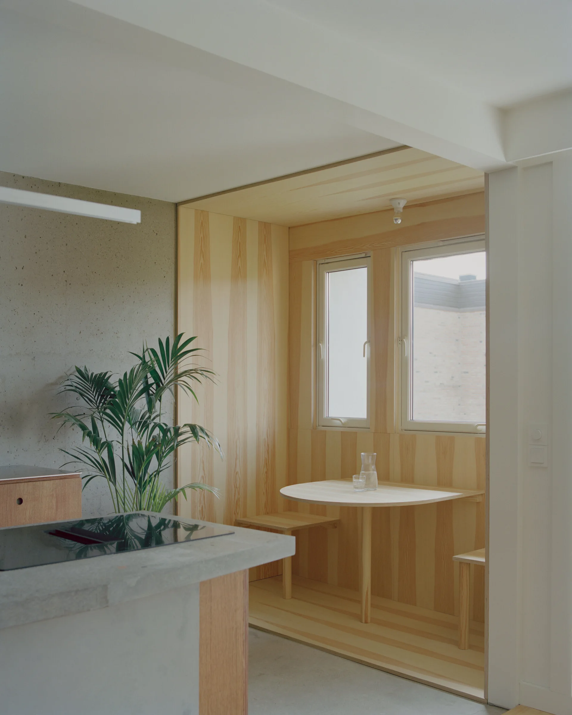 A modern kitchen featuring a concrete island in the foreground and a built-in light wood dining nook with a small circular table and bench seating.
