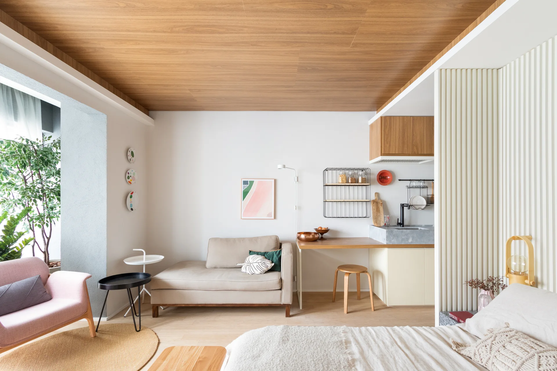 A modern studio apartment features a warm wood ceiling, a beige sofa, and a white fluted partition wall separating the kitchen and sleeping areas.