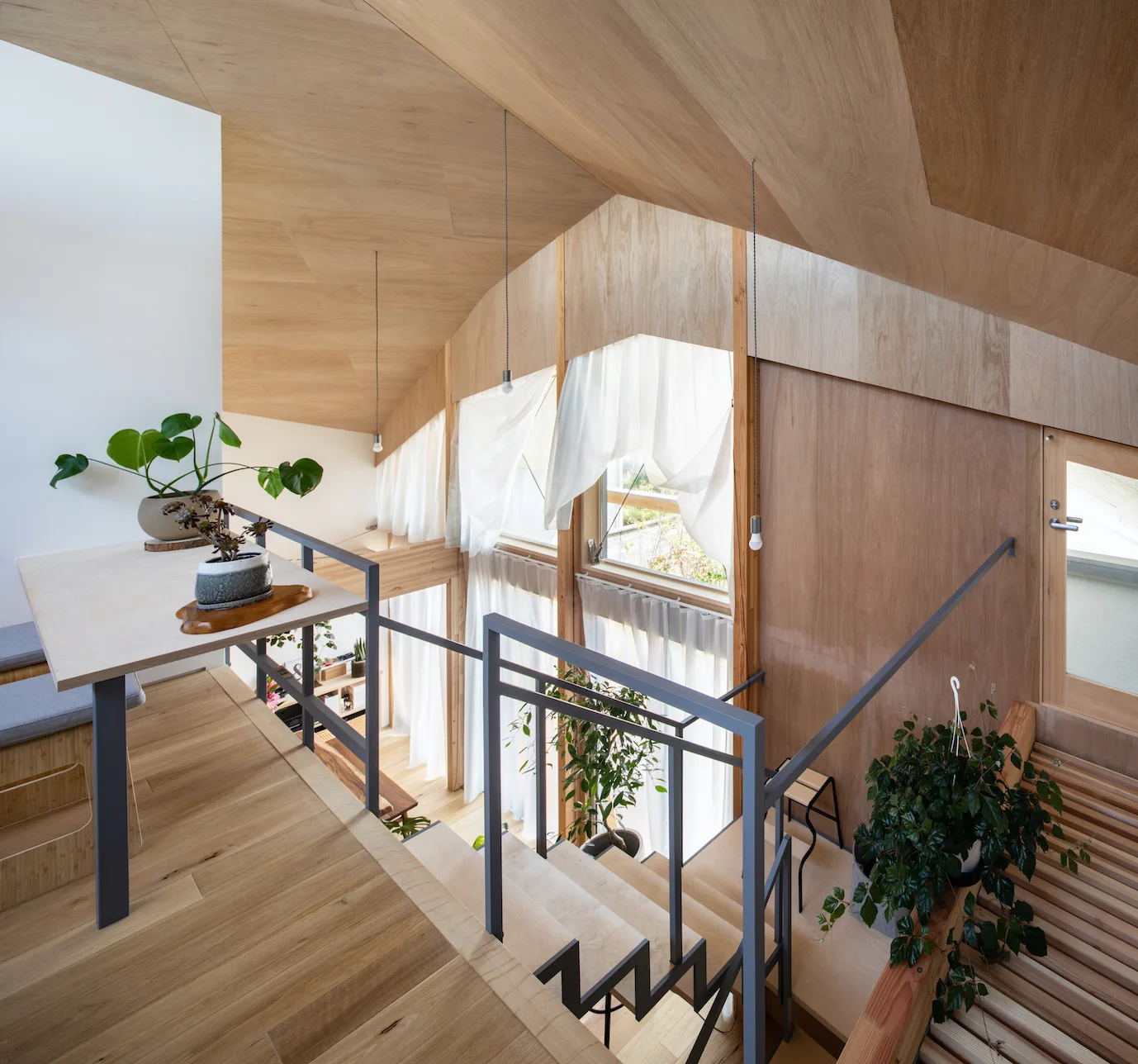 A light wood staircase leads up to a loft landing with a sloped plywood ceiling and large windows draped in sheer curtains.