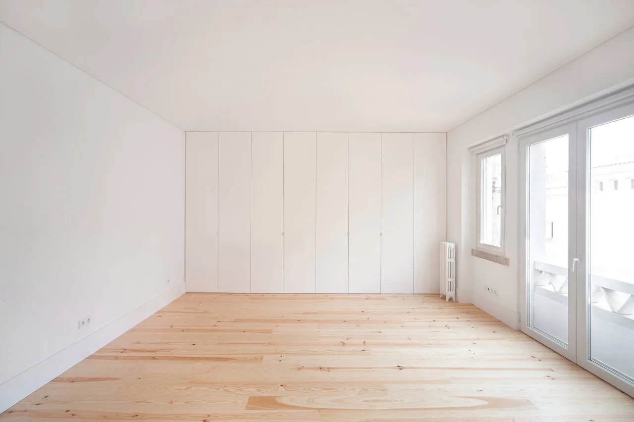 An empty minimalist room featuring natural light wood plank floors, clean white walls, and a full wall of floor-to-ceiling built-in white cabinetry.