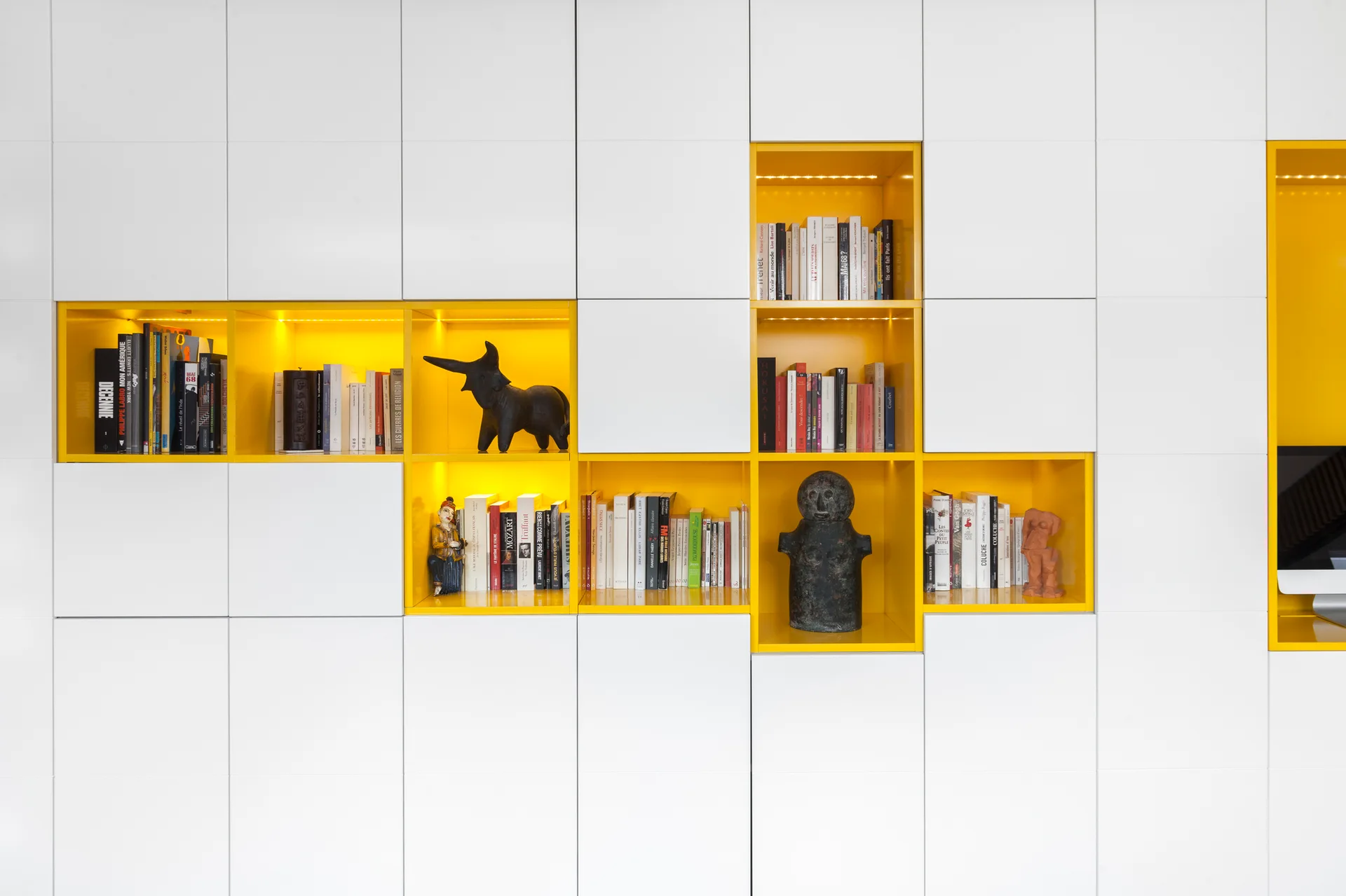 A white modular storage wall featuring asymmetrical yellow-lined cabinet niches illuminated with LED strips and filled with books and small sculptures.