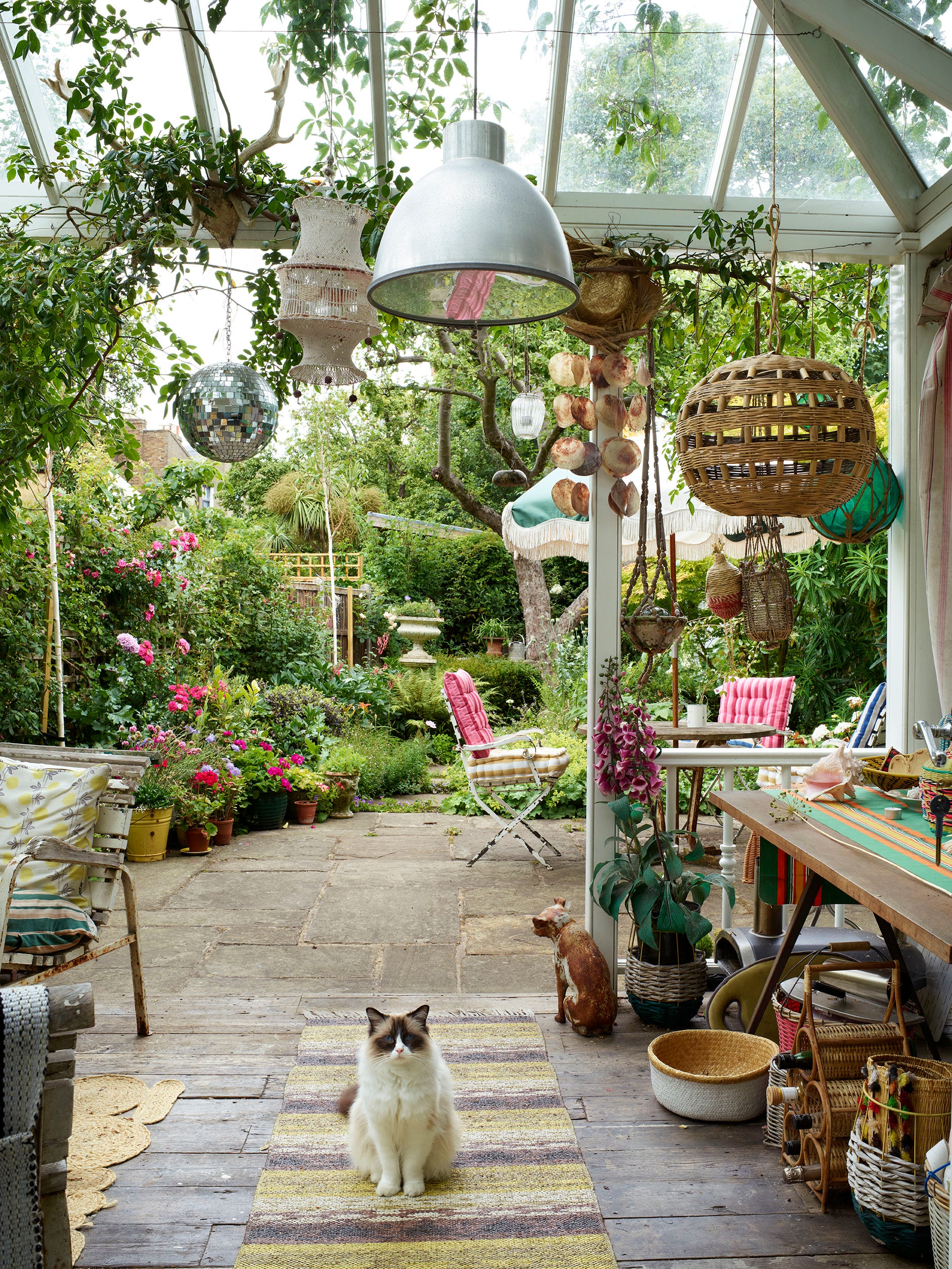 A sunlit glass-roofed patio features a wooden table, hanging wicker and metal pendant lights, potted plants, and a cat sitting on a striped rug.