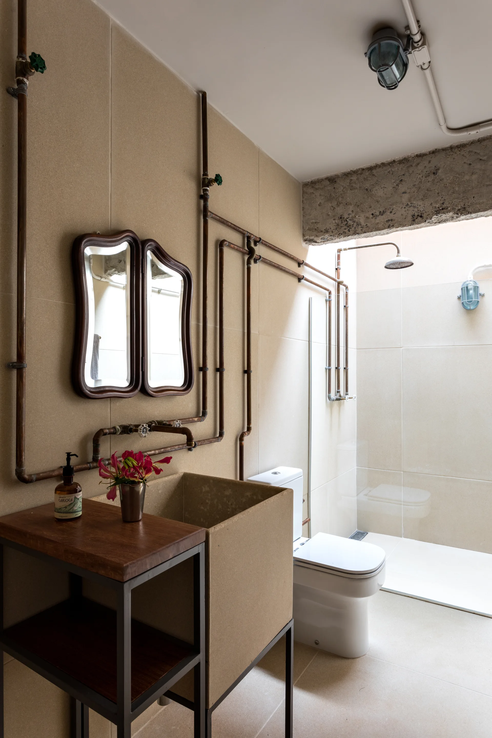 An industrial bathroom features exposed copper plumbing pipes on beige tiled walls, a cast concrete block sink, and wavy dark wood mirrors.