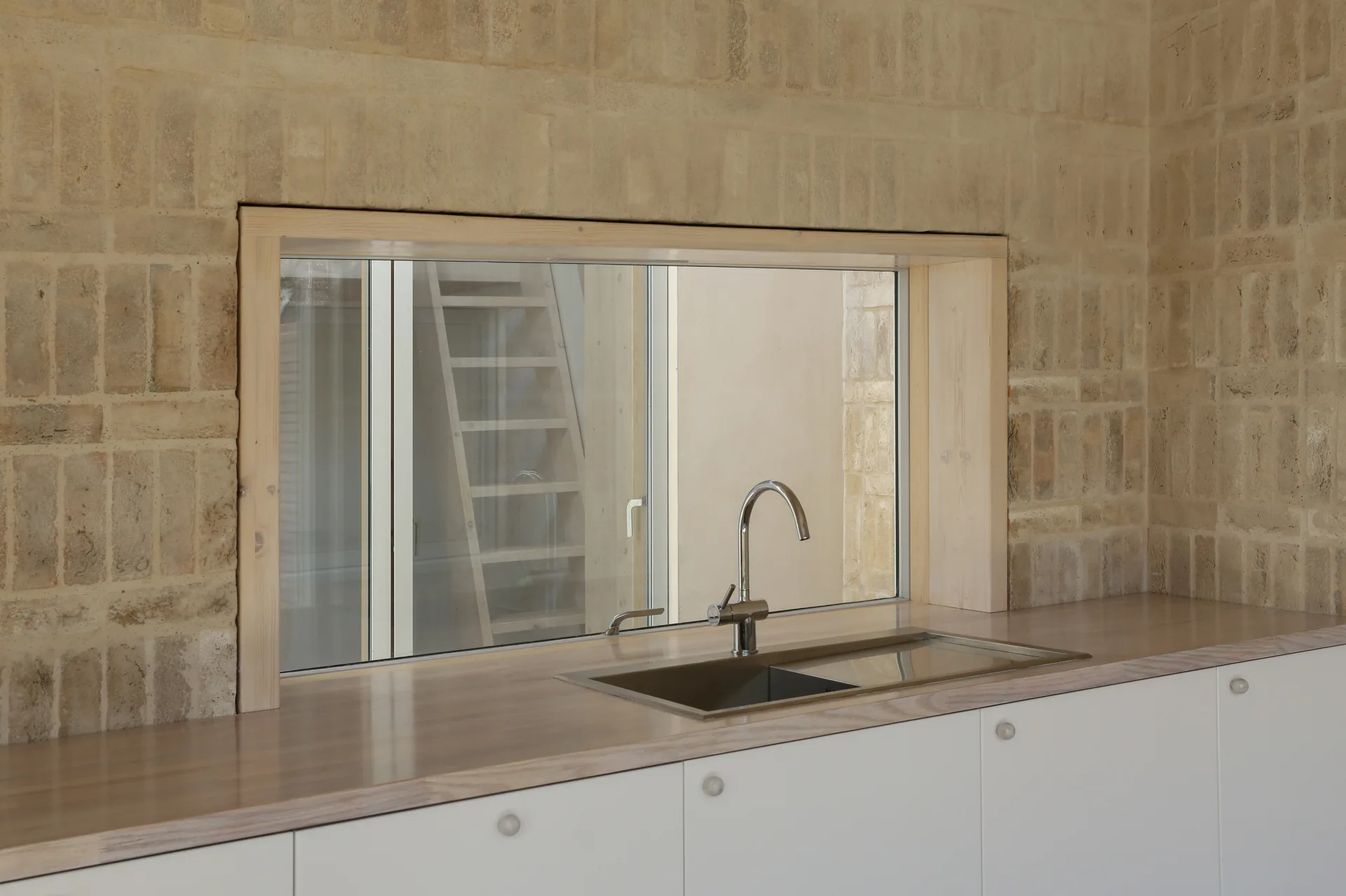 A stainless steel kitchen sink set into a light wood countertop against a textured beige brick wall with a large wood-framed window.