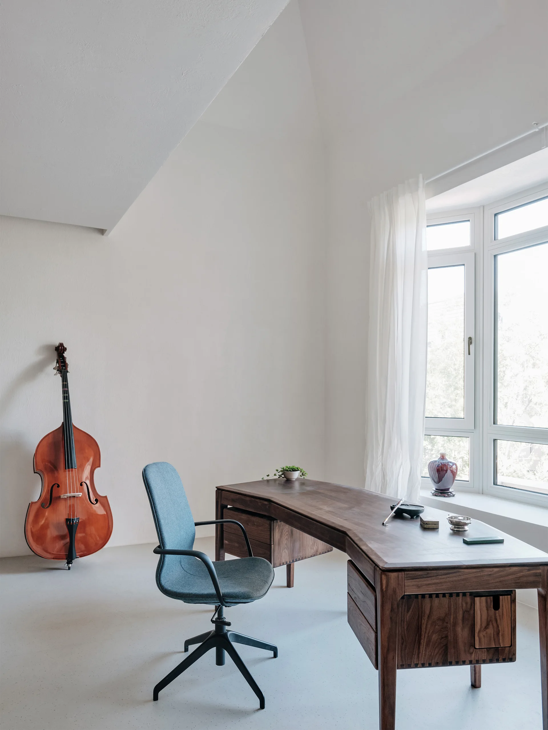 A bright minimalist home office features a curved dark wood desk, a blue office chair, and an upright bass leaning against a white wall.