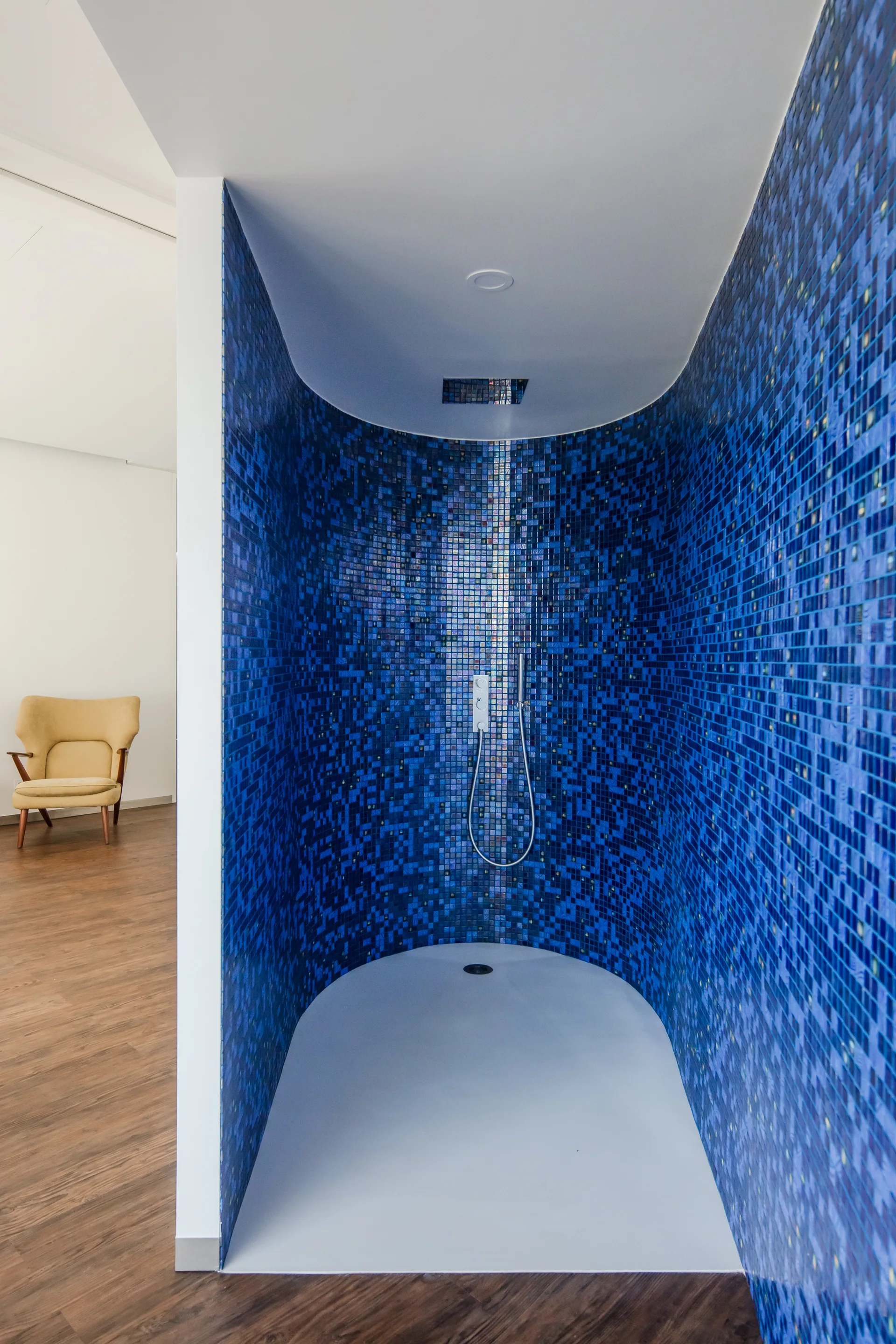 A curved walk-in shower lined with blue mosaic tile stands next to dark wood flooring and a yellow armchair.