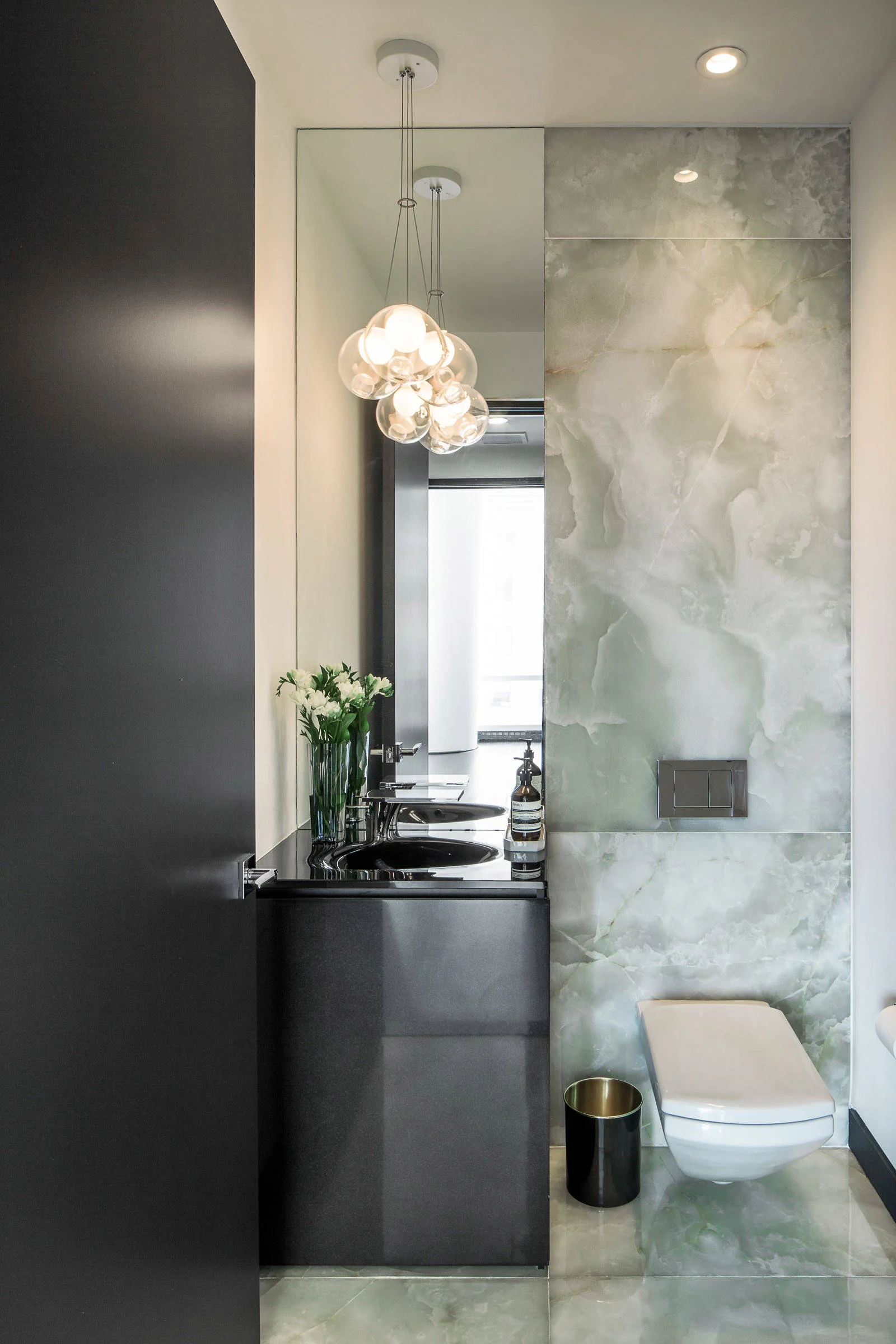 A modern powder room with a black vanity, a wall-mounted toilet, green veined stone wall paneling, and a hanging cluster of glass globe pendant lights.