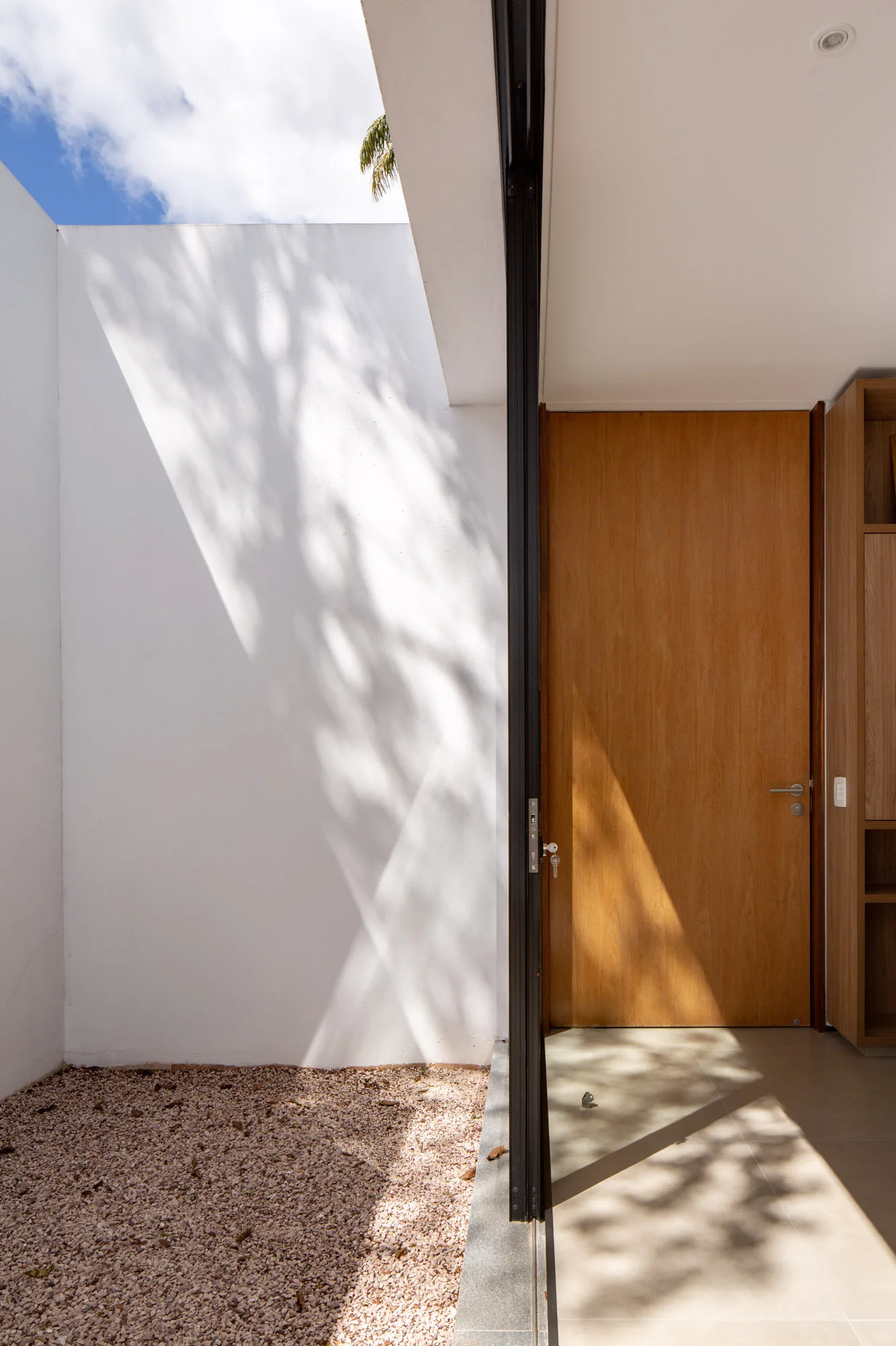 A minimalist interior entryway with a wooden door opens to a white-walled courtyard with gravel flooring and tree shadow patterns.