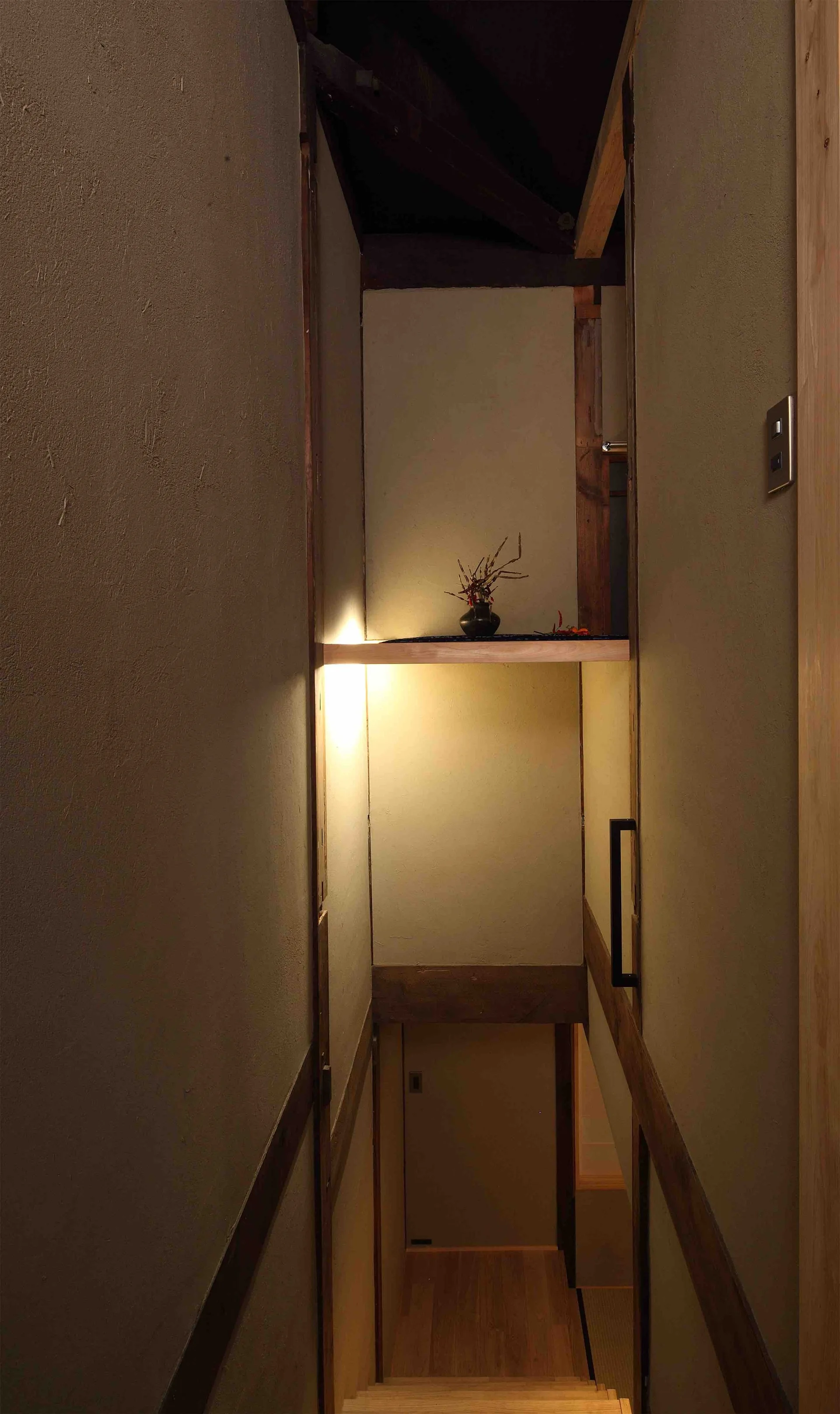 A narrow staircase with textured plaster walls features an illuminated recessed wooden shelf displaying a small dark vase with branches.