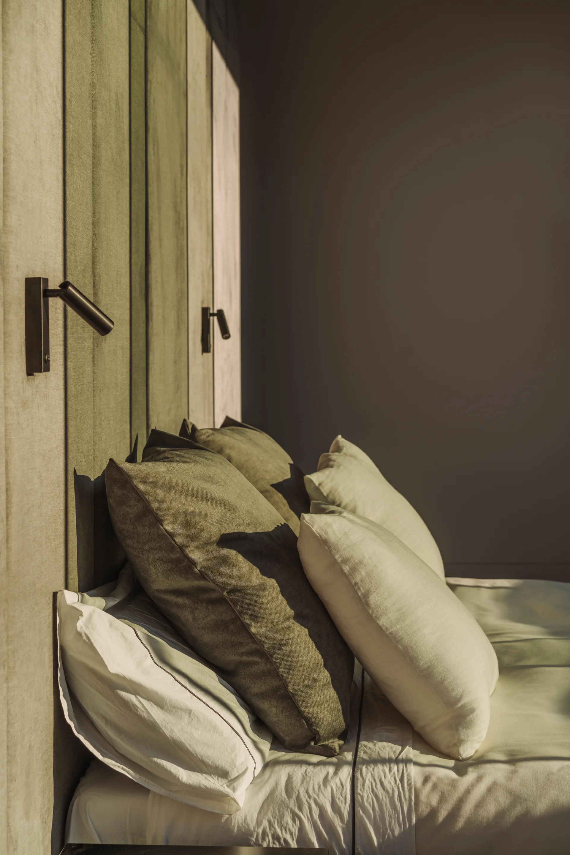 An olive green upholstered paneled headboard wall features mounted black reading lights behind layered pillows and white bed sheets.