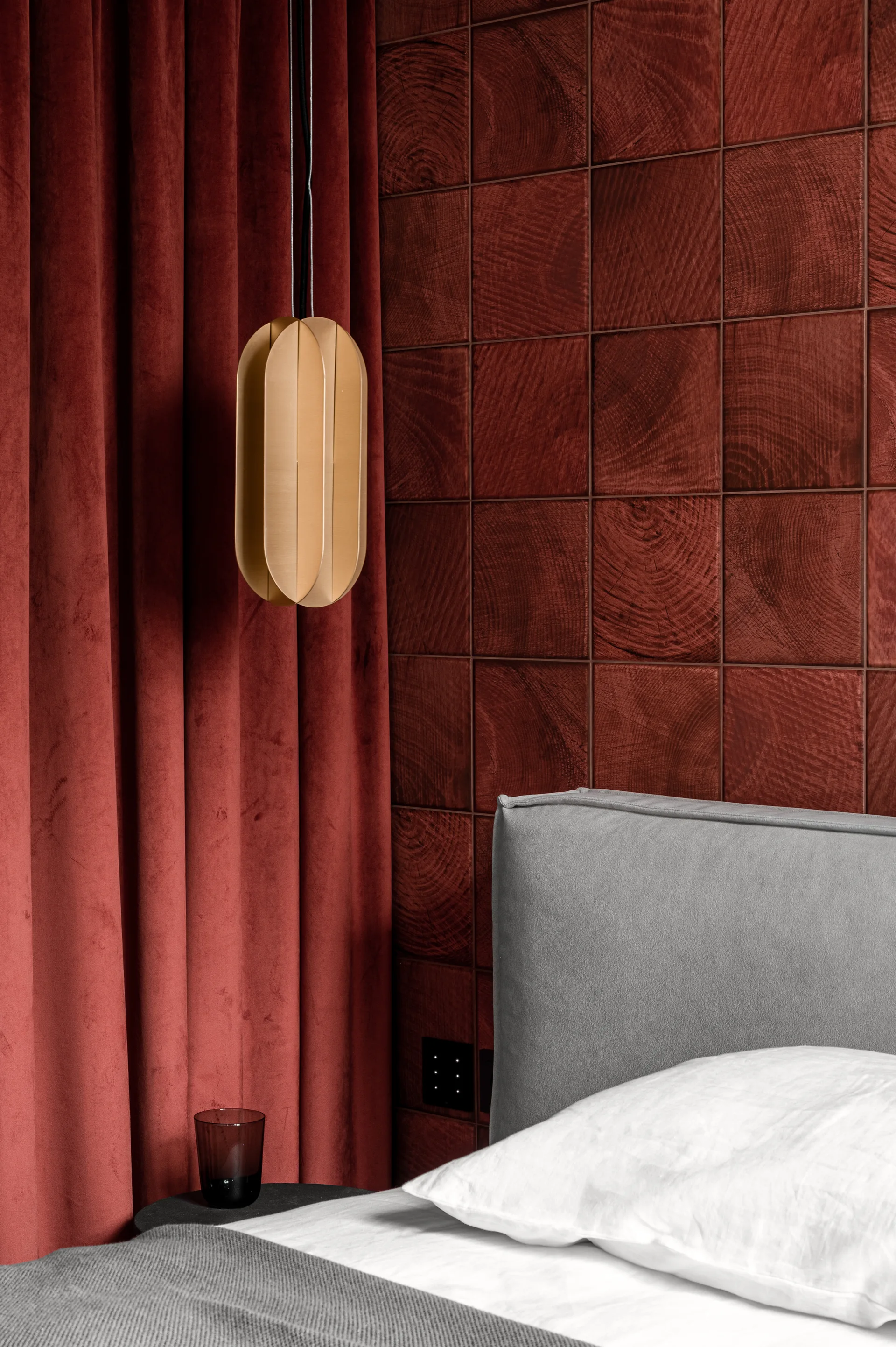 A bedroom corner features a grey upholstered headboard set against a red wood tile wall, beside matching velvet curtains and a brass pendant light.