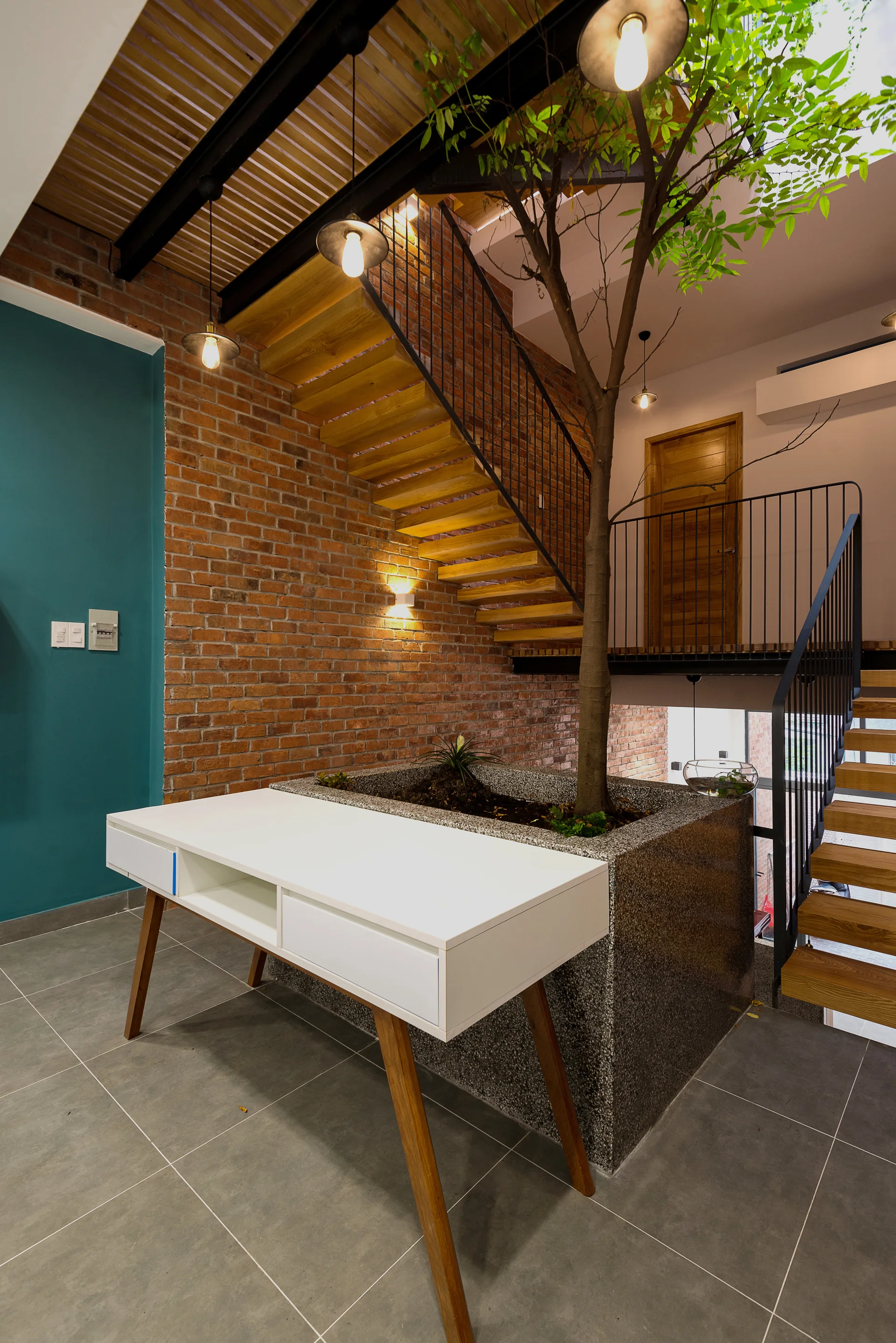 A modern wooden staircase rises along an exposed brick wall, featuring a large textured planter with an indoor tree and a white desk in the foreground.