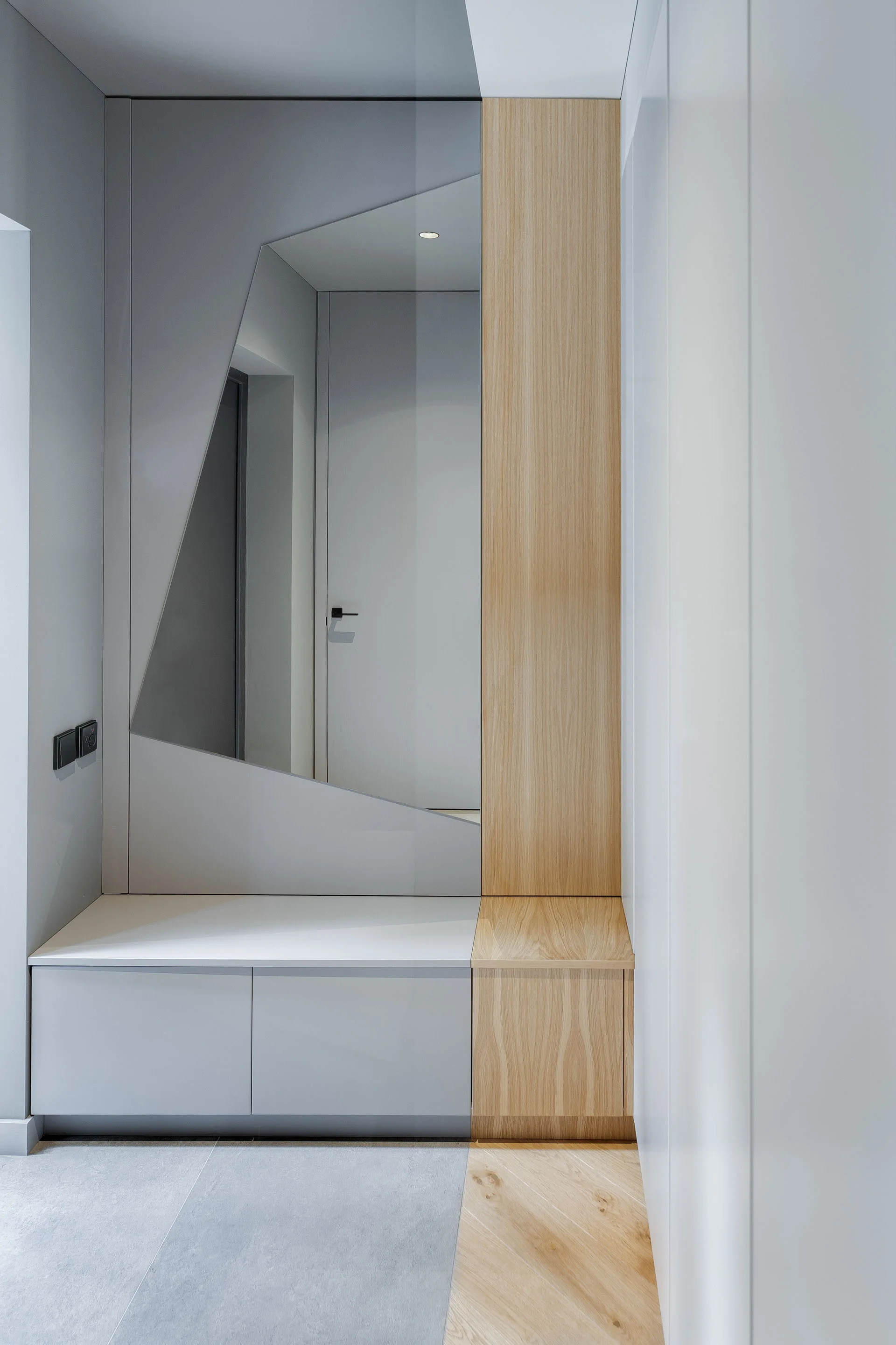 A minimalist entryway featuring built-in grey cabinetry integrated with an asymmetrical mirror, a wood-toned storage seat, and matching diagonal flooring.
