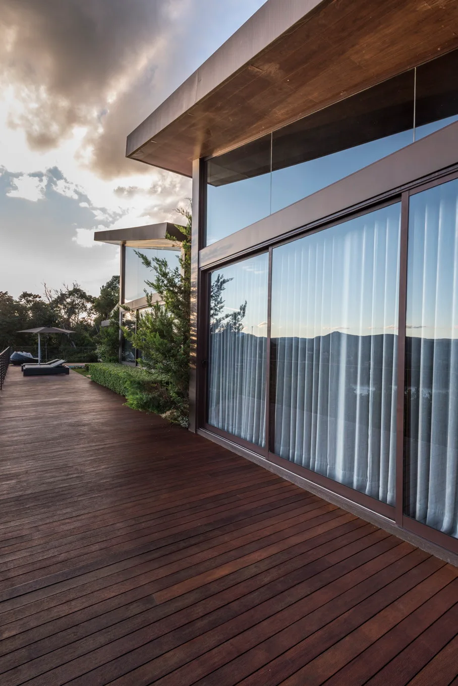 A spacious wood-plank deck extends along a modern home with large glass sliding doors and white sheer curtains.