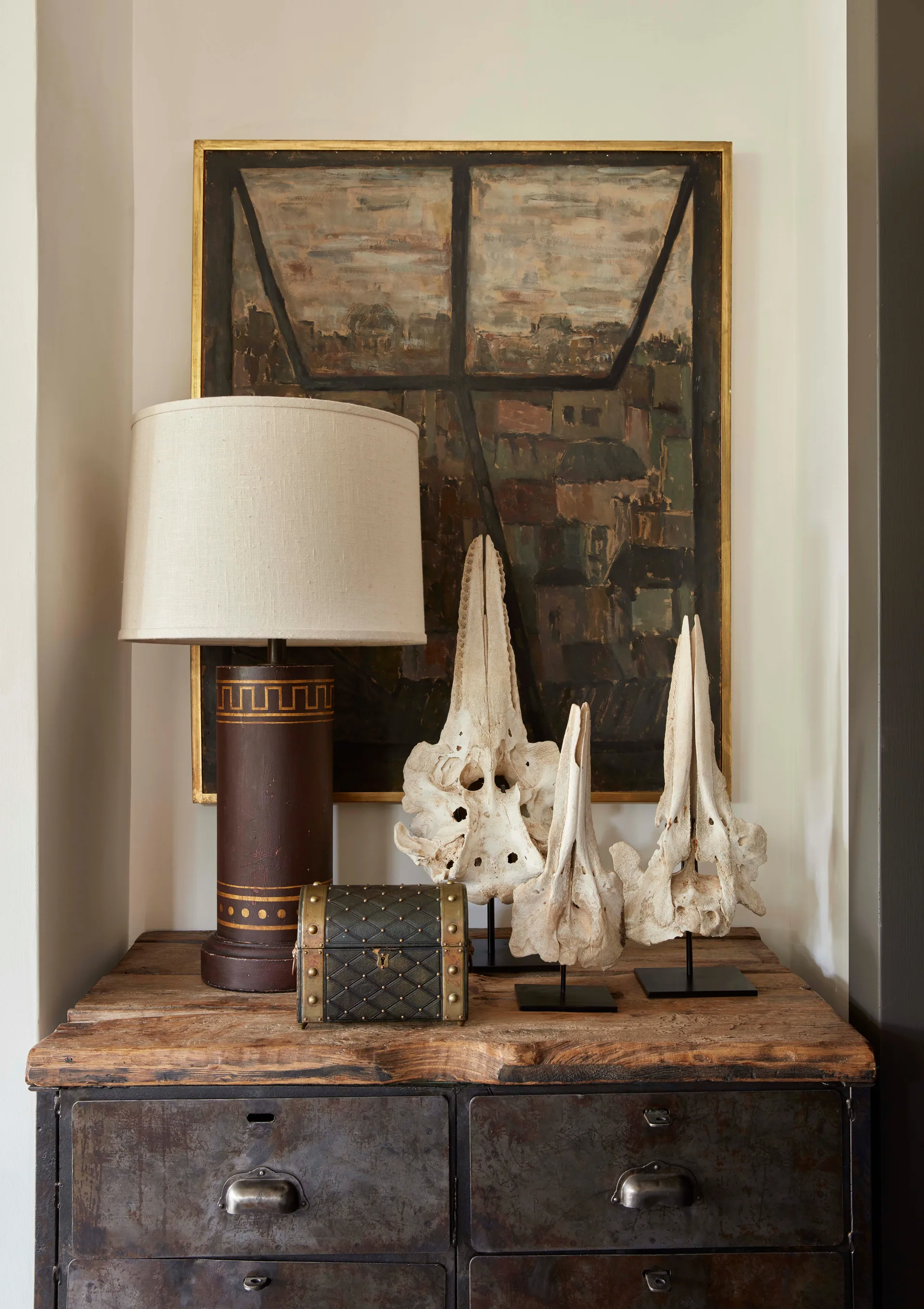 Three bone sculptures on stands, a brown table lamp, and a small chest sit on a weathered wood and metal industrial dresser in front of a framed painting.