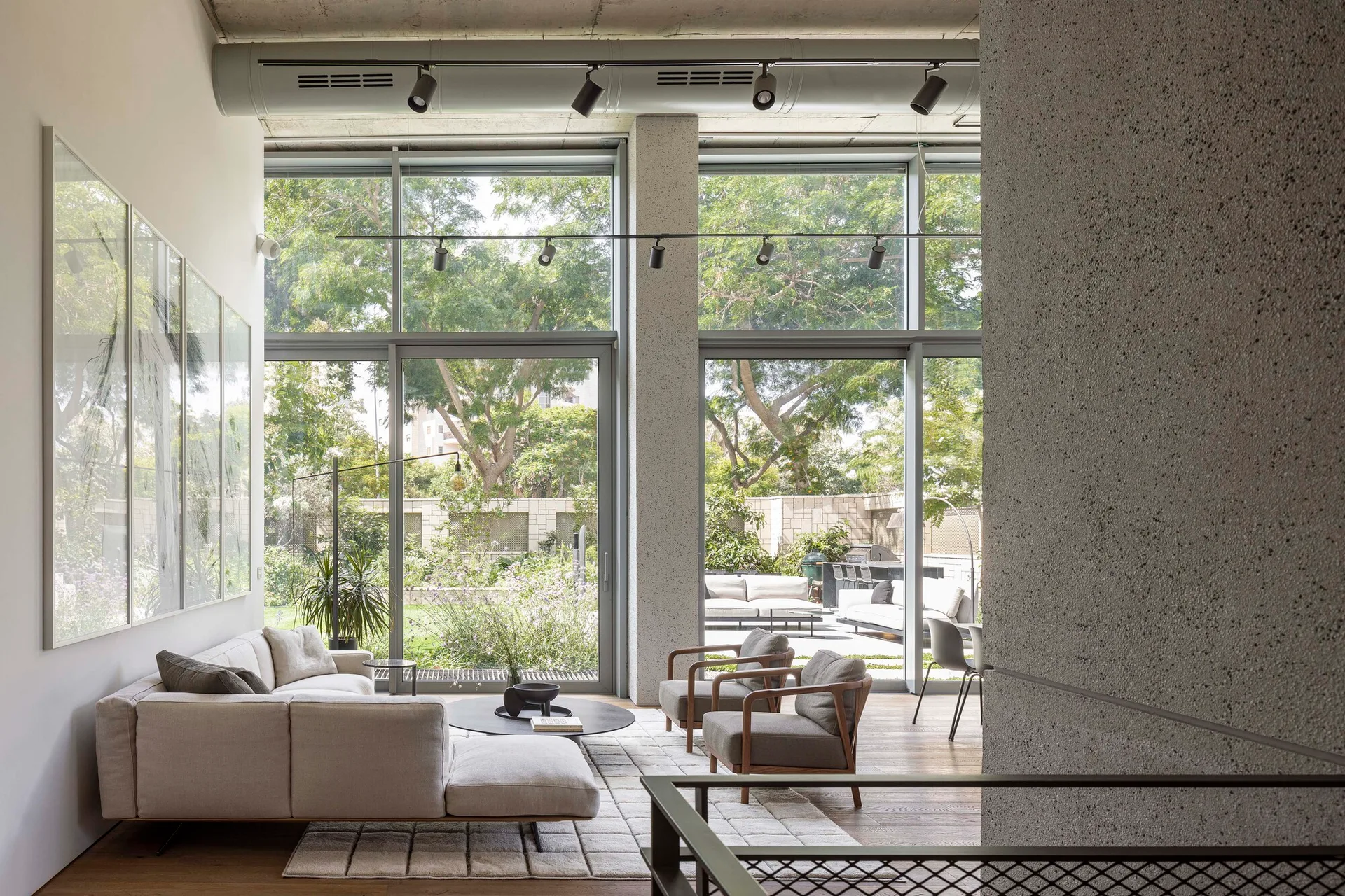A modern living room features a cream sectional sofa, wooden armchairs, and a textured rug under an exposed concrete ceiling with floor-to-ceiling windows.