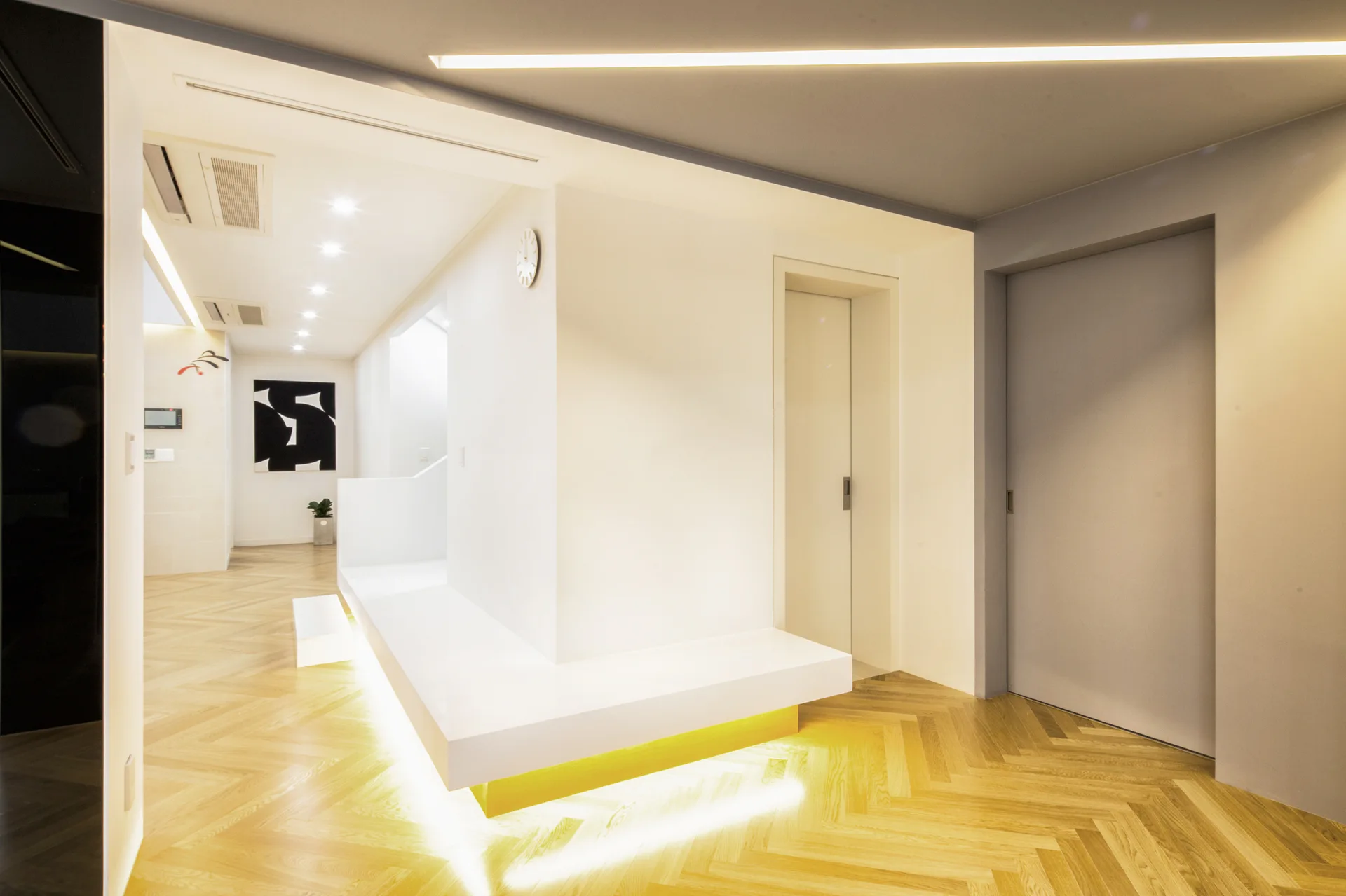 A modern minimalist hallway features light wood herringbone flooring, a white cantilevered bench with warm under-lighting, and clean white walls.