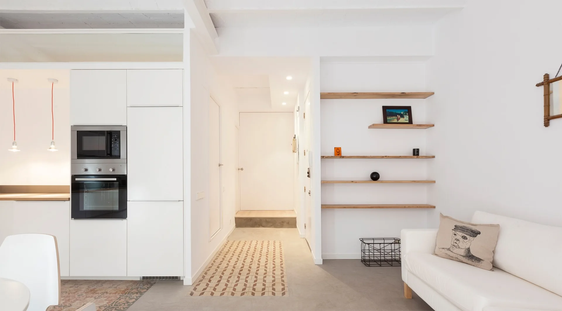 A minimalist white apartment interior featuring integrated kitchen cabinetry, a geometric-tiled hallway path, wooden open shelves, and a white sofa.