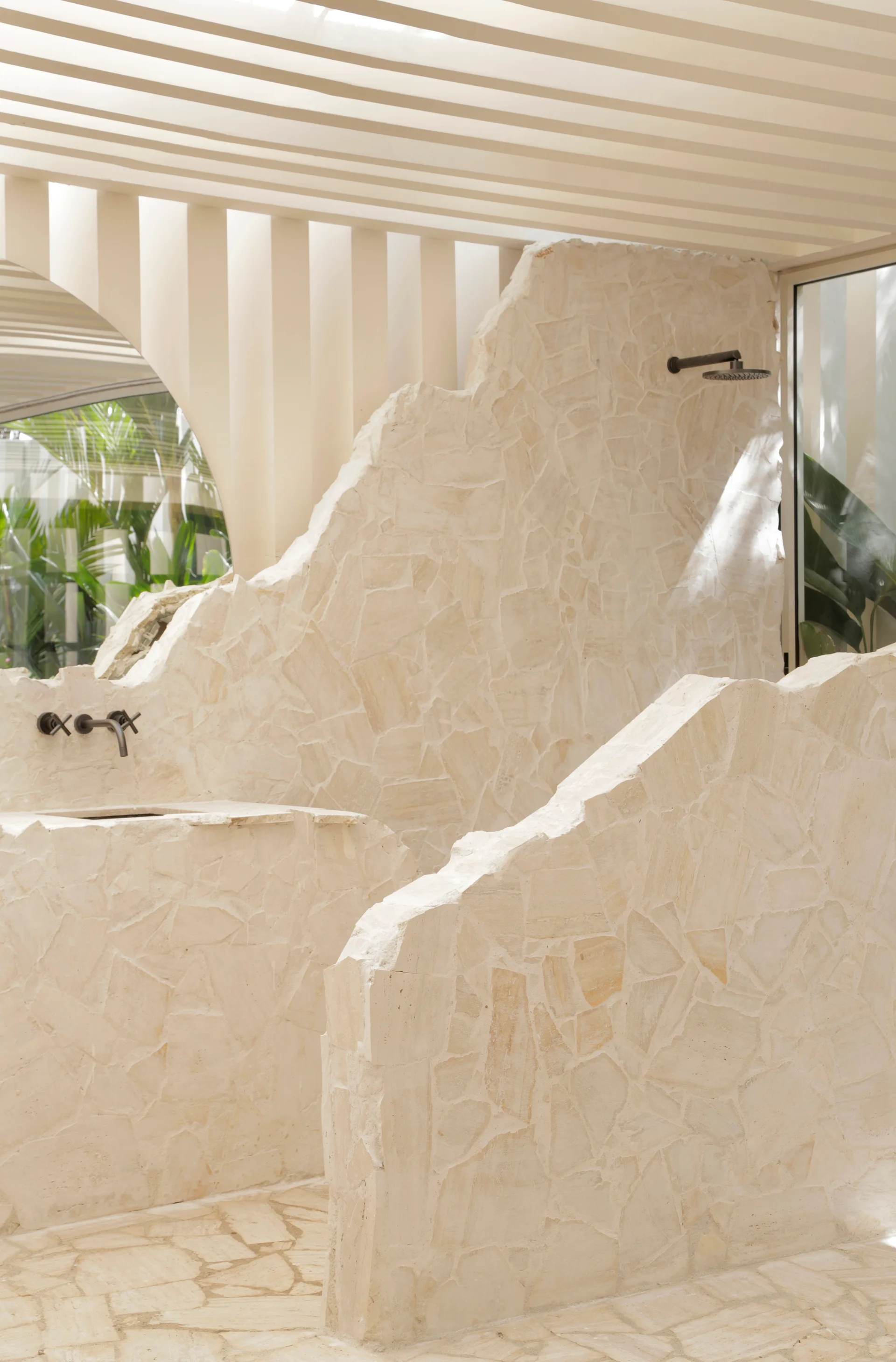 A beige stone bathroom features jagged-edge partition walls, a built-in sink with a dark faucet, and a shower under a white slatted ceiling.