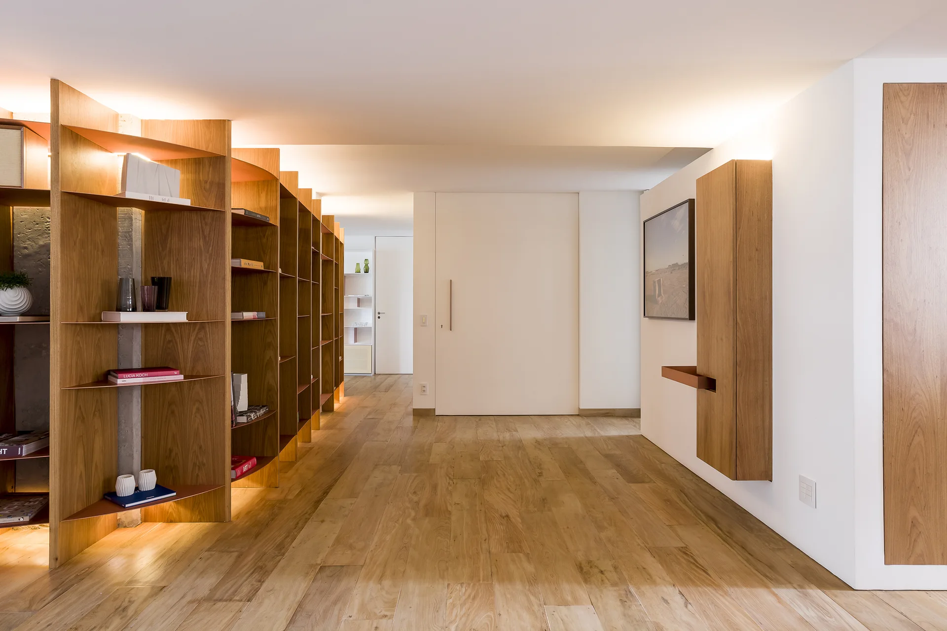 A modern hallway features custom wood shelving with integrated lighting, light wood floors, and a large white sliding door.