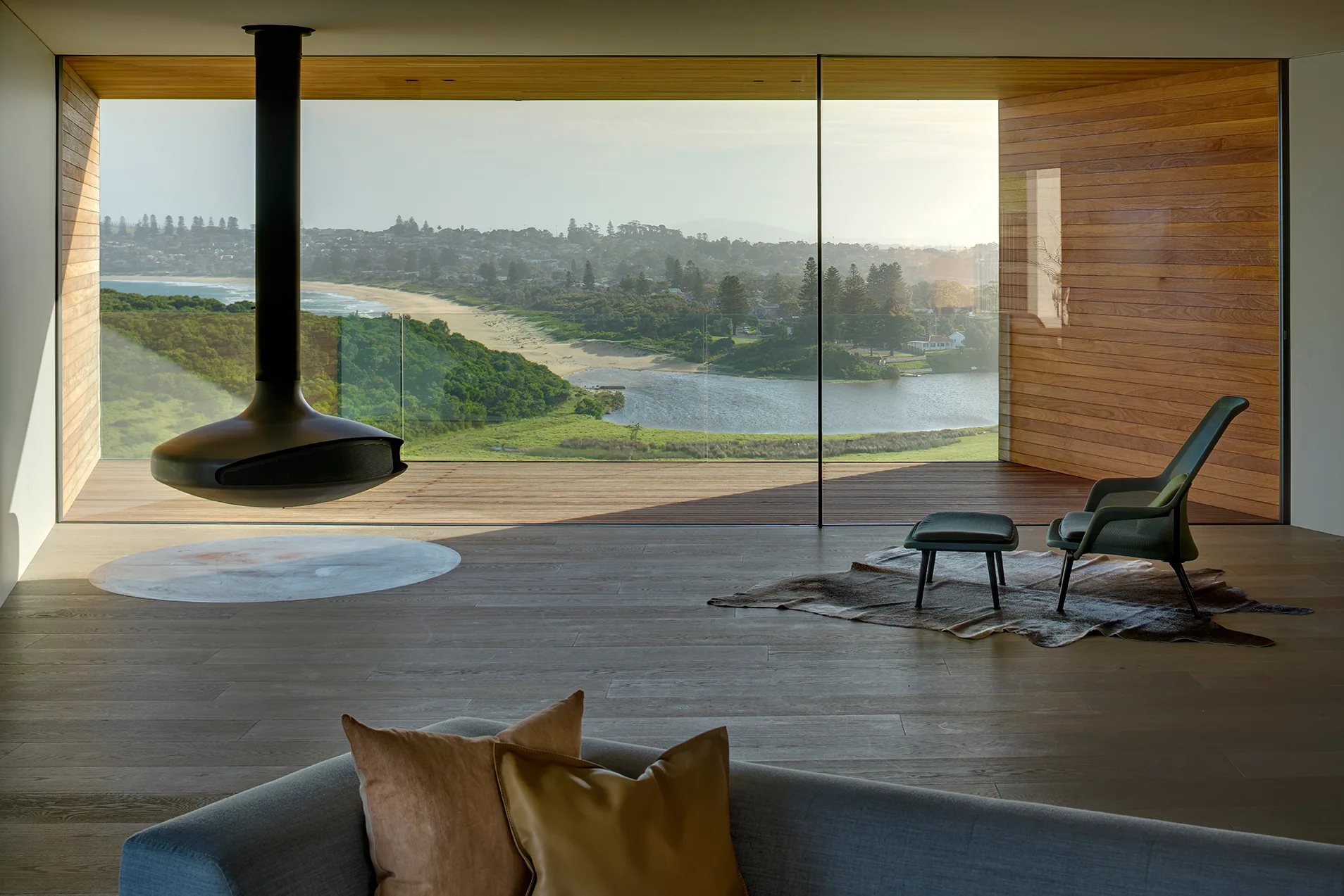 A modern living room features a black suspended fireplace, a green armchair, and an animal hide rug in front of large glass walls overlooking a coastline.