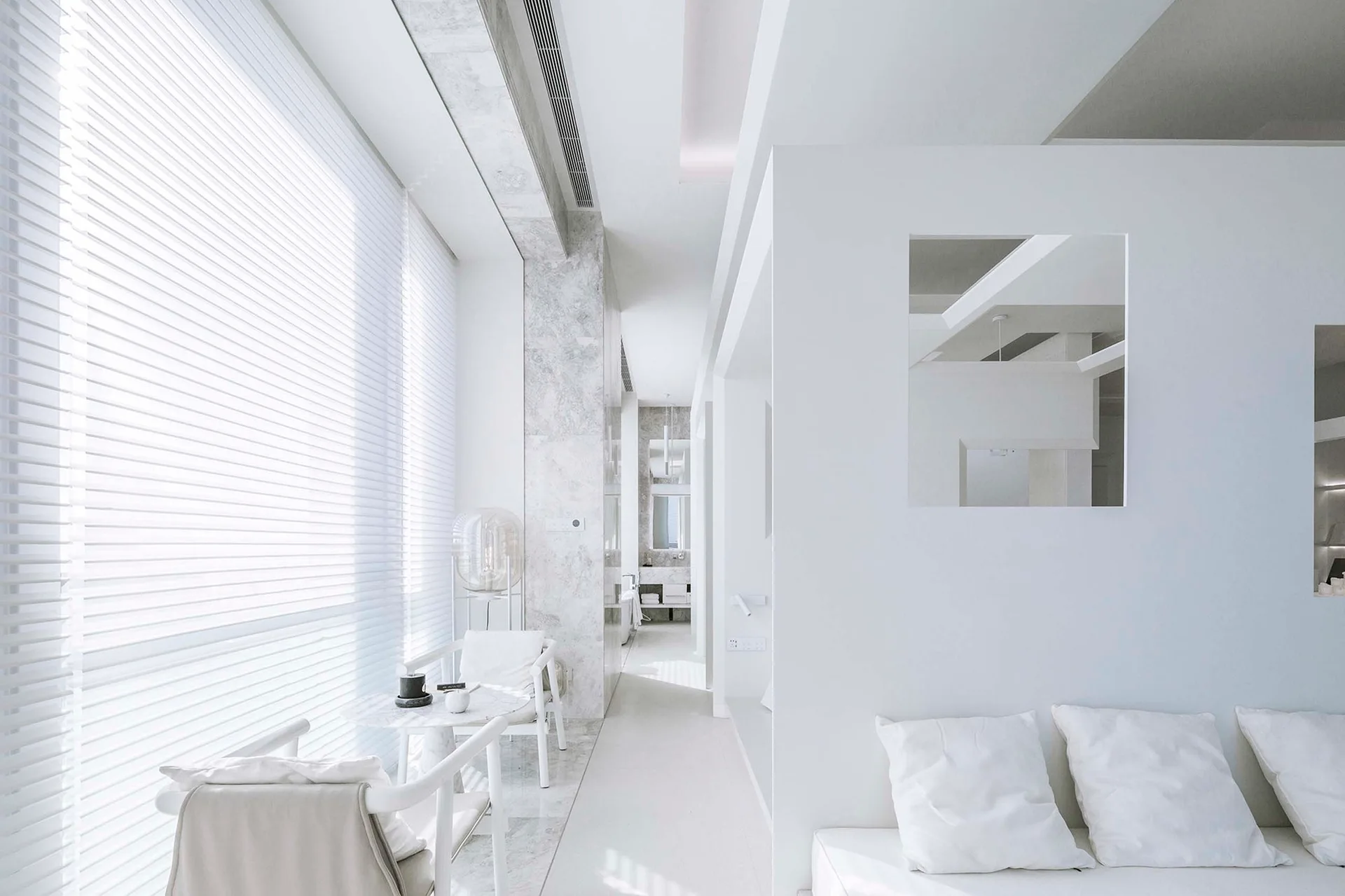 A minimalist white living area with horizontal window blinds, a small marble-top bistro table, and a square wall cutout.