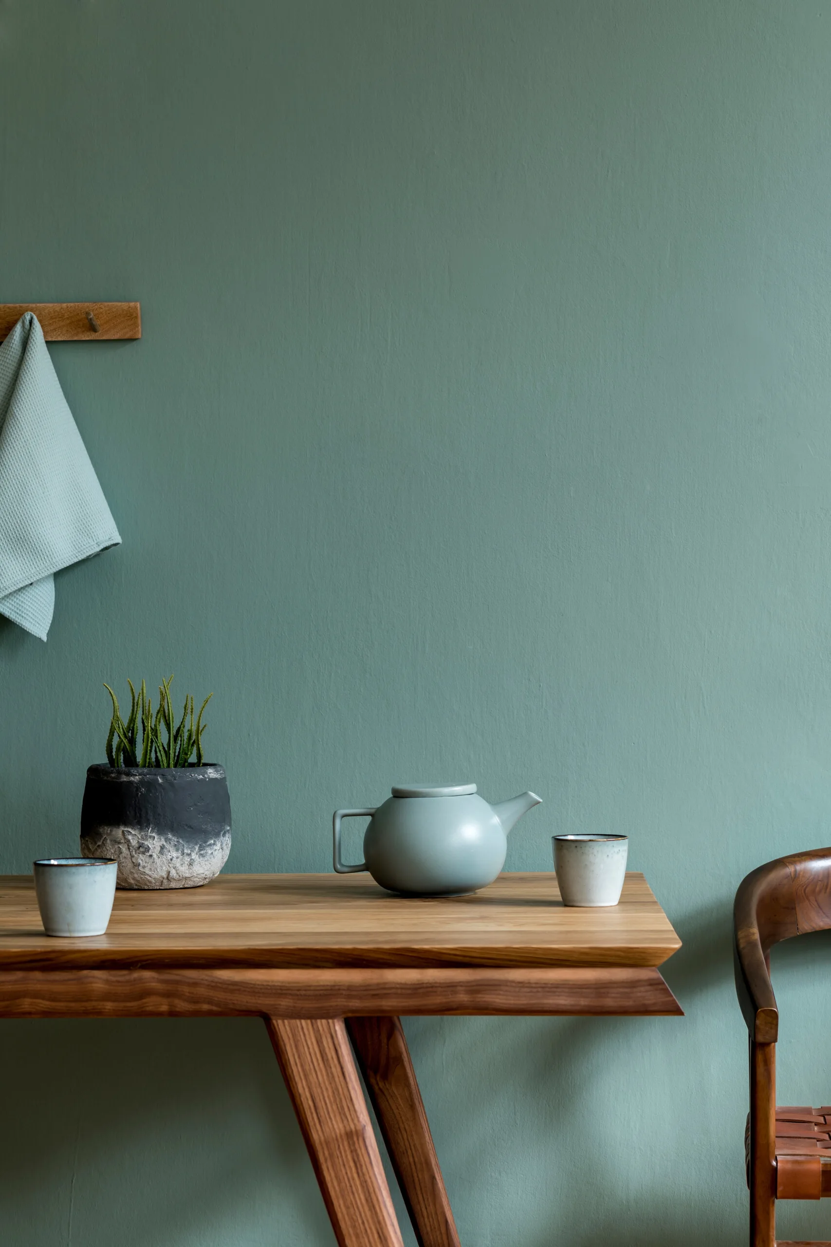 A wooden dining table set with a sage green teapot and small ceramic cups stands against a matching sage green wall in a minimalist room.