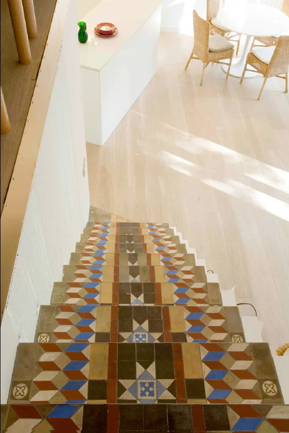 A top-down view of a staircase featuring colorful geometric tiles leading to a bright dining space with light wood floors and wicker chairs.