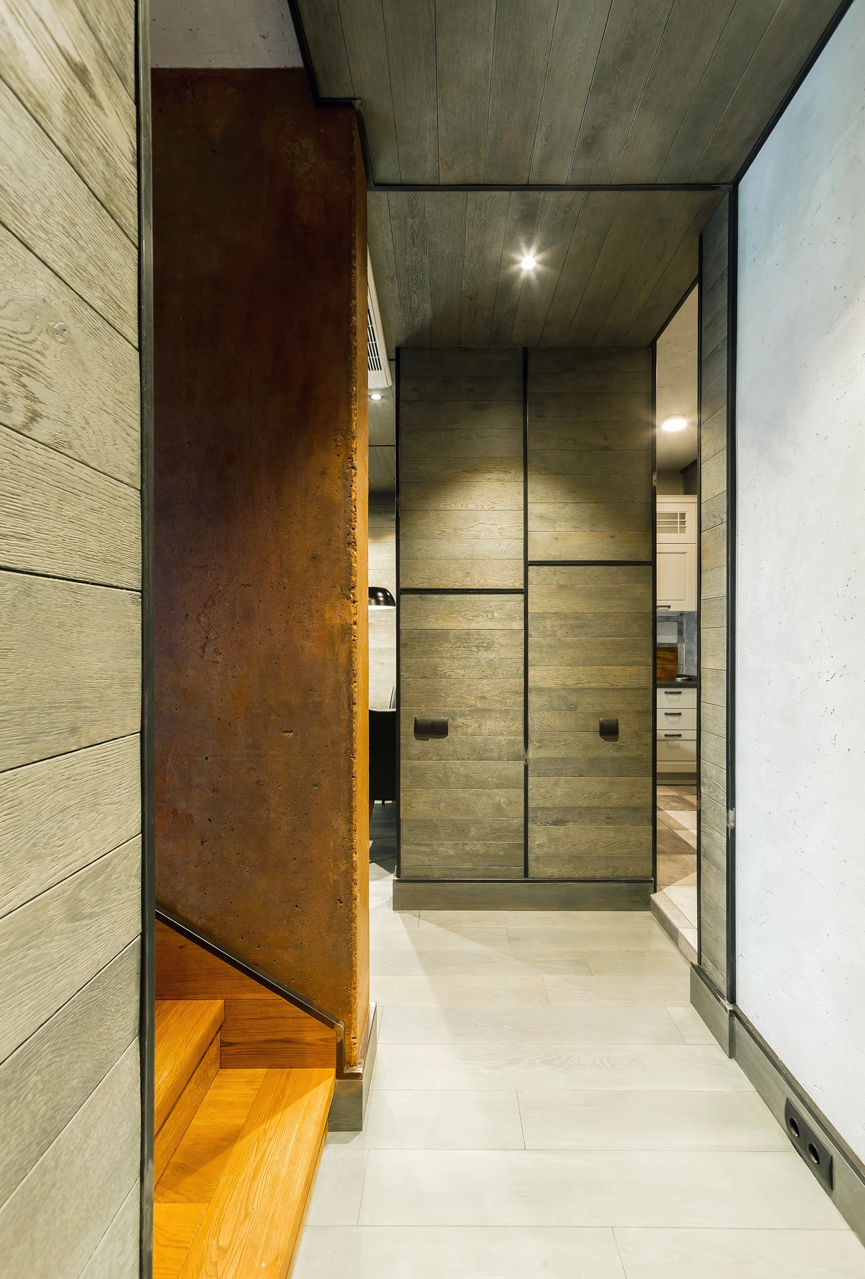 A modern hallway features horizontal grey wood wall paneling, a rust-brown textured column, light wood stairs, and built-in matching storage cabinets.