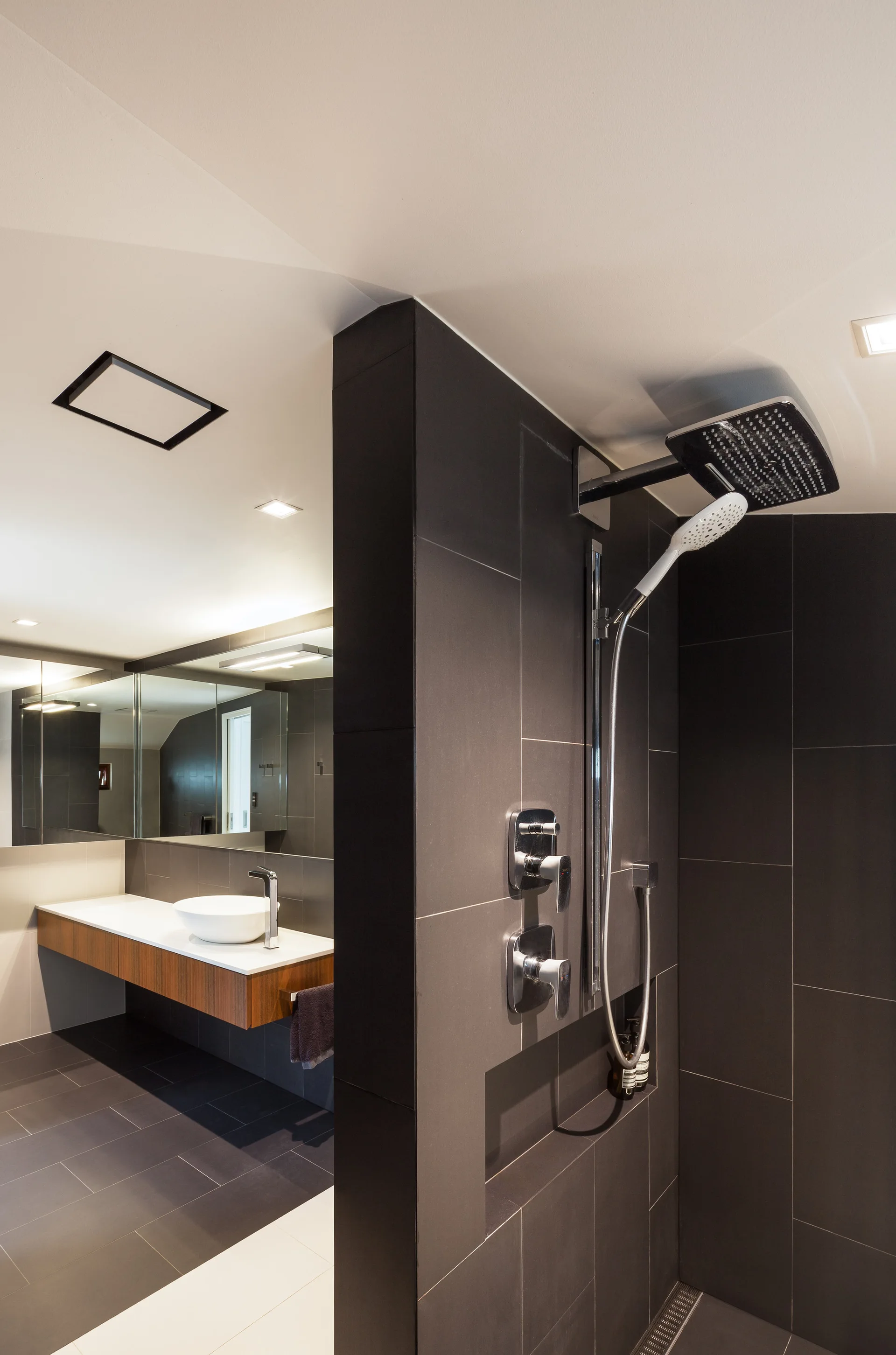A modern bathroom features a dark charcoal tile shower partition next to a wall-mounted wooden vanity with a white vessel sink.