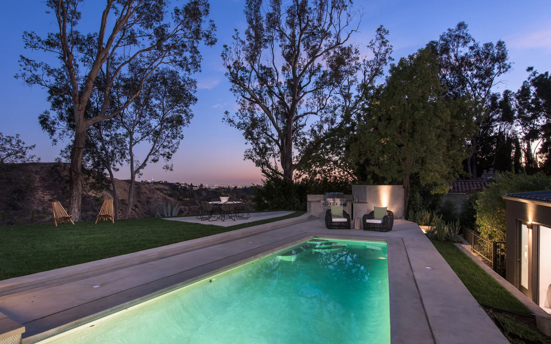 An illuminated rectangular pool glows green next to a concrete patio, built-in outdoor kitchen, and dining area at twilight.